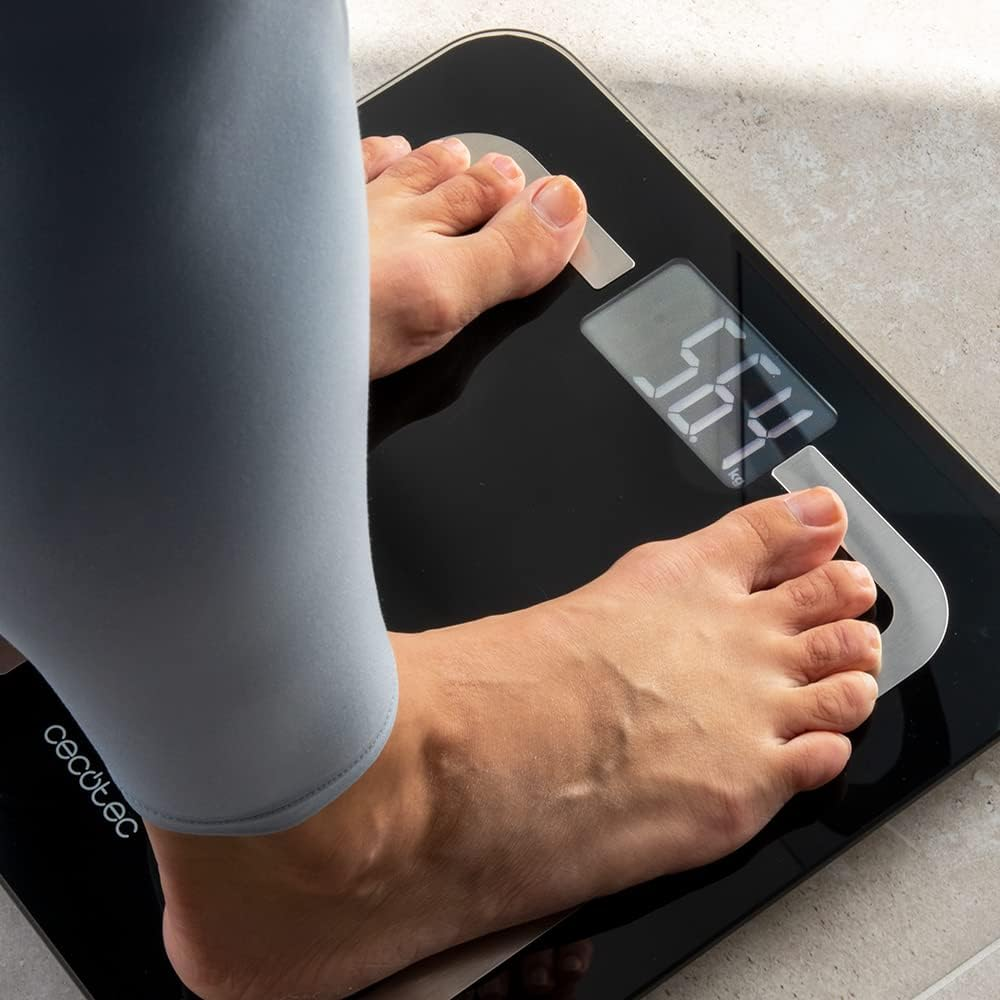 Persona de pie en una báscula digital negra, mostrando 56.4 kg. Piernas con leggings grises, pies en la báscula, en un suelo embaldosado.