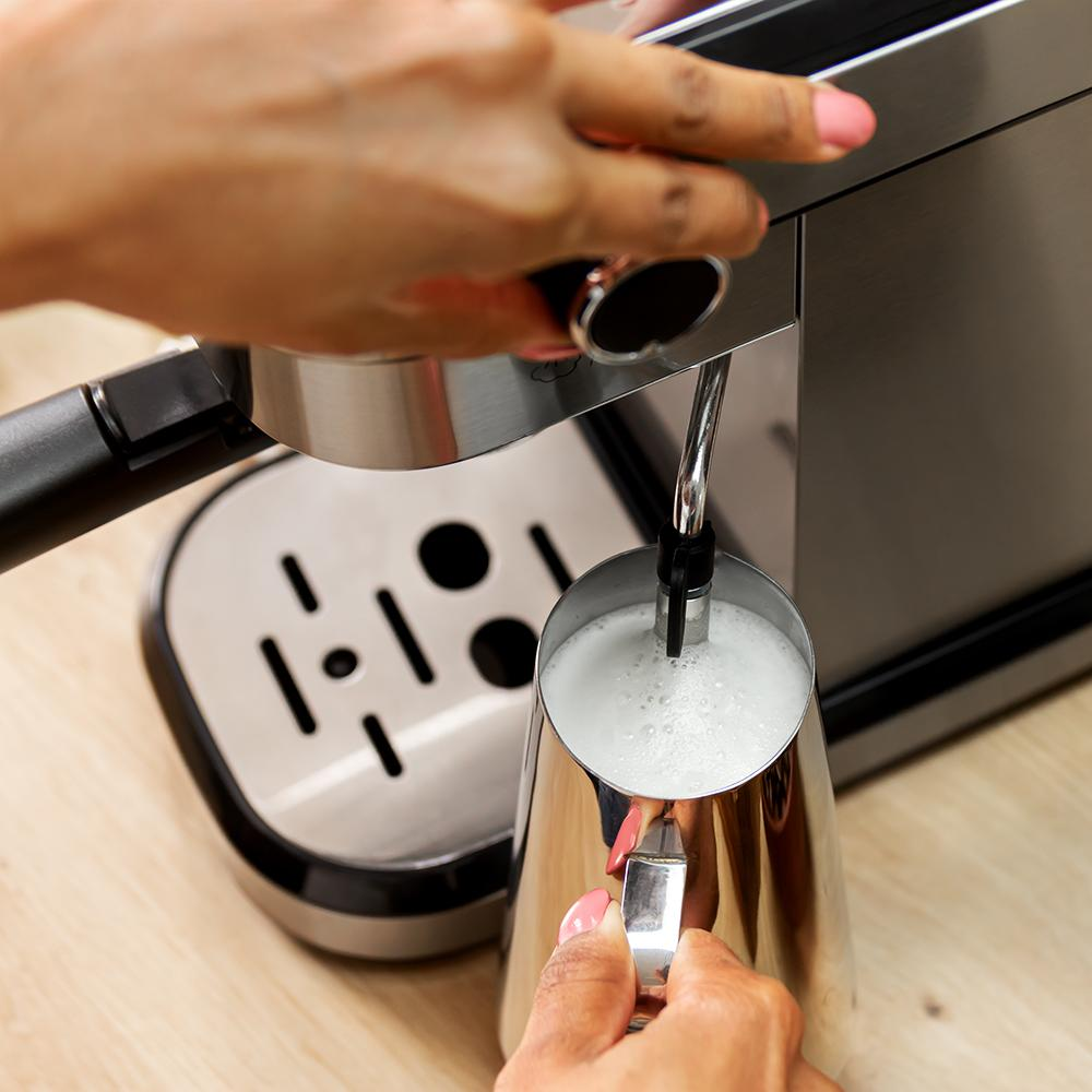 Manos operan una máquina de espresso, espumando leche en una jarra de metal. La máquina es plateada y negra, sobre una superficie de madera.