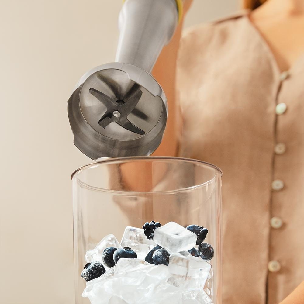 Una batidora está sobre un vaso con cubitos de hielo y arándanos. Una mujer con una camisa beige está en el fondo.
