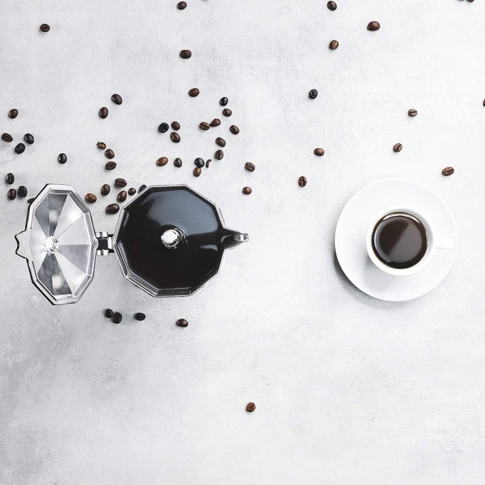 Vista aérea de granos de café esparcidos alrededor de una cafetera y una taza de café sobre una superficie blanca.
