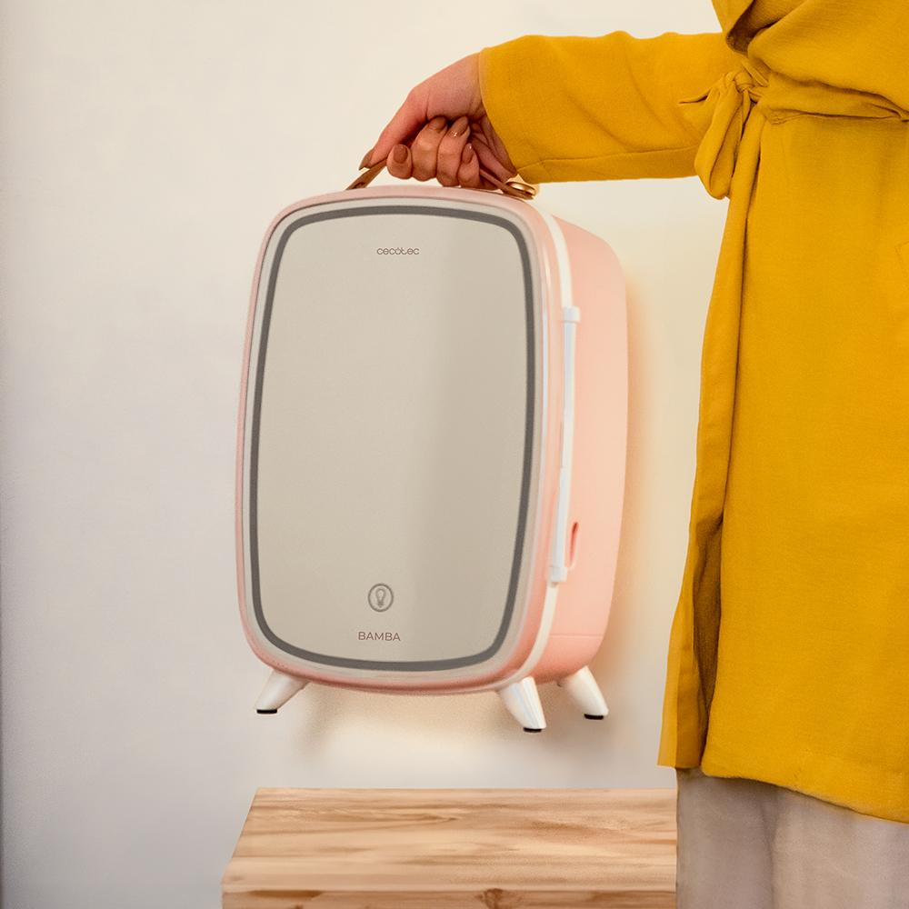 Una persona sostiene una mini nevera rosa contra una pared blanca. La nevera tiene una puerta de espejo y la persona lleva una chaqueta amarilla.
