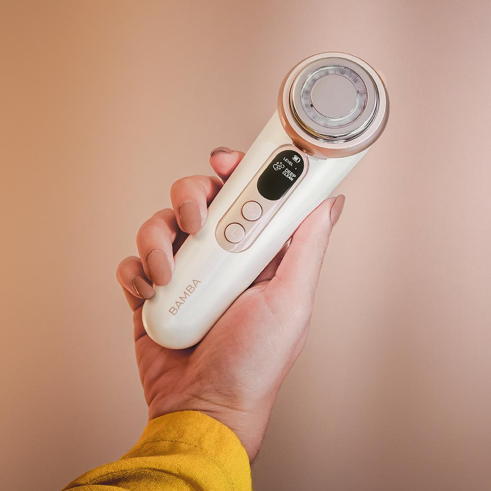 Una mano sostiene un dispositivo blanco para el cuidado de la piel. El dispositivo tiene una parte superior metálica y botones. El fondo es melocotón.