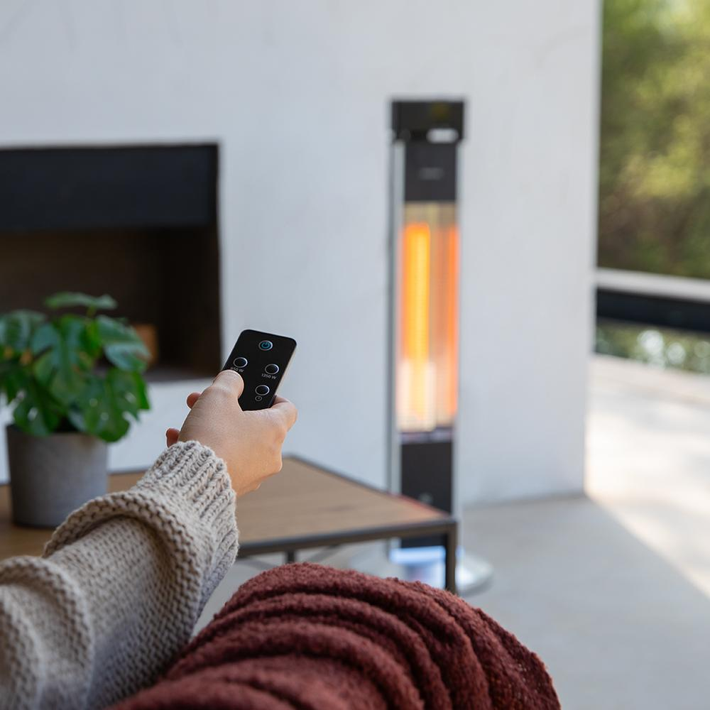 Una persona sostiene un control remoto, operando un calefactor de patio. El calefactor está encendido, con un brillo naranja brillante. Una planta en maceta es visible.