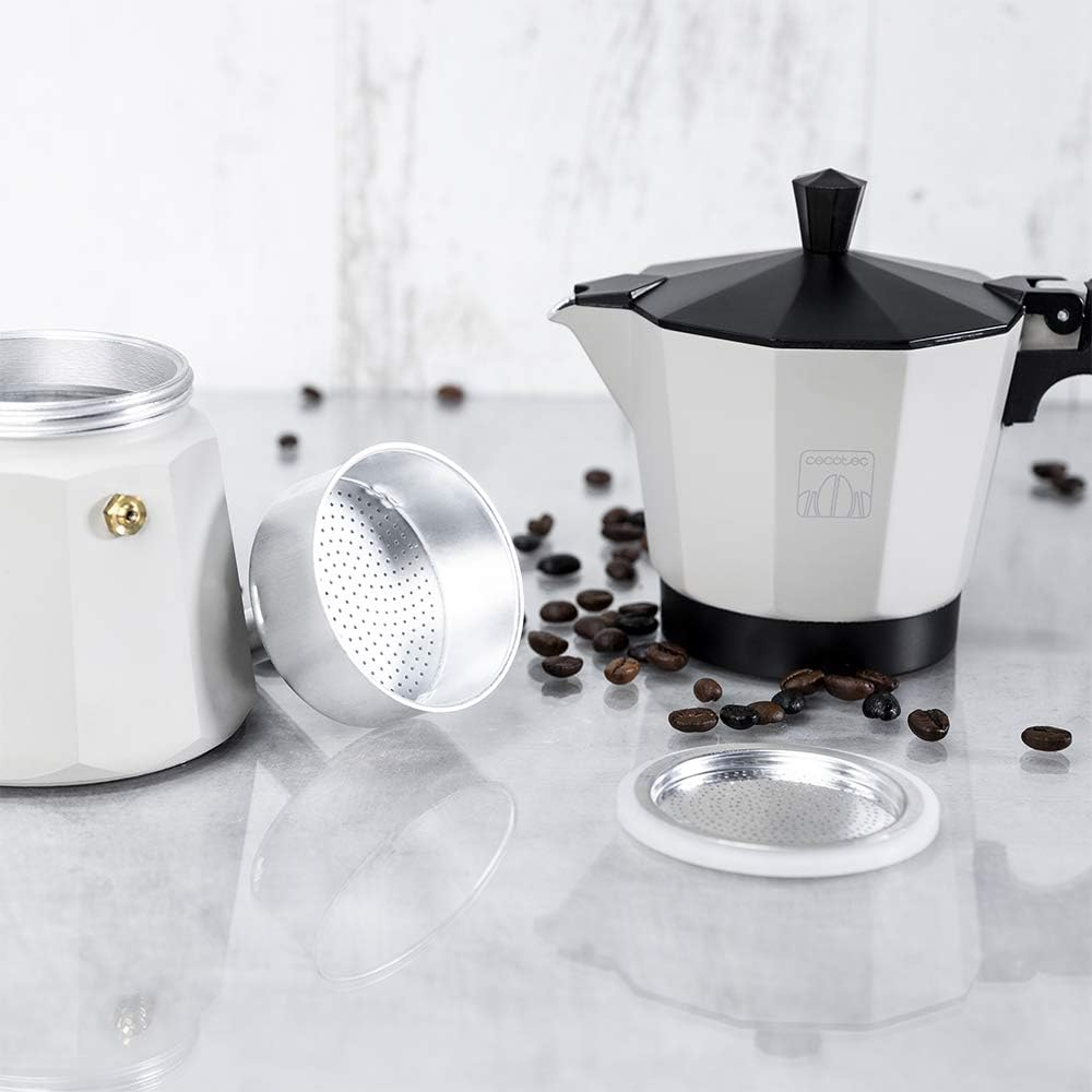 Una cafetera desmontada con granos de café esparcidos. La cafetera es blanca y negra. Componentes y granos están sobre una superficie reflectante.