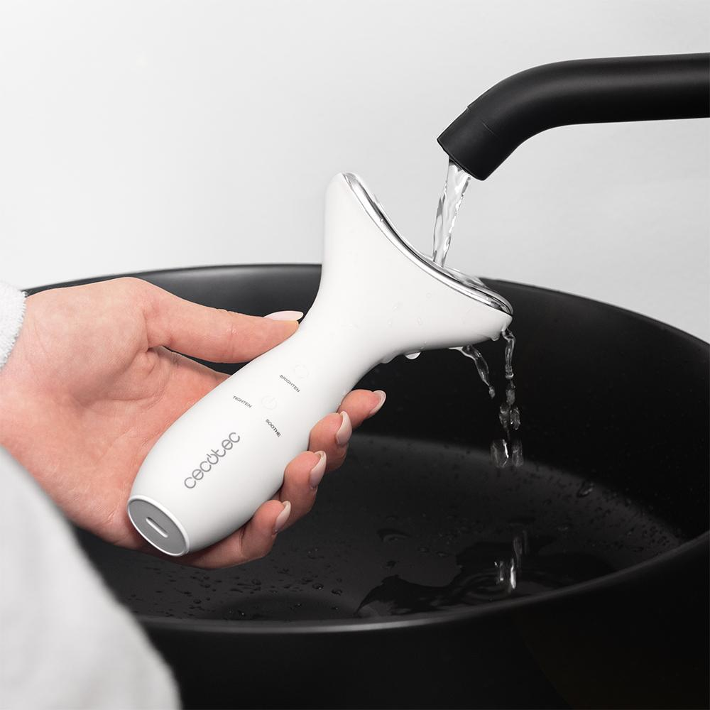 Un masajeador facial blanco es sostenido por una mano. El agua fluye de un grifo negro sobre el masajeador y en un cuenco negro.