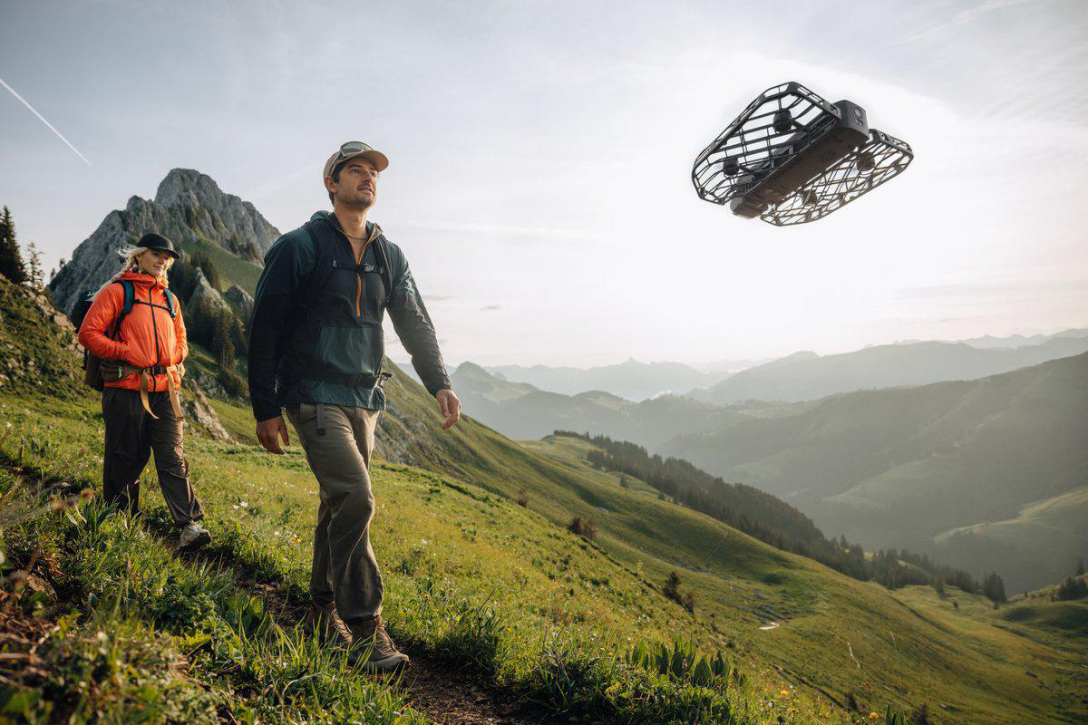 Zwei Wanderer auf einem grünen Hügel mit einem Berg im Hintergrund. Eine Drohne fliegt über ihnen. Der Himmel ist klar.