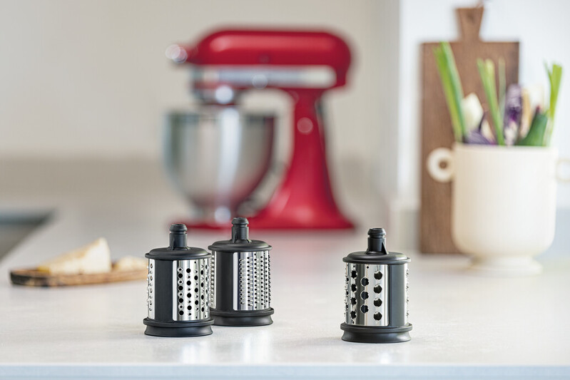 Three black and silver kitchen graters on a white counter. A red stand mixer and vegetables are in the background.
