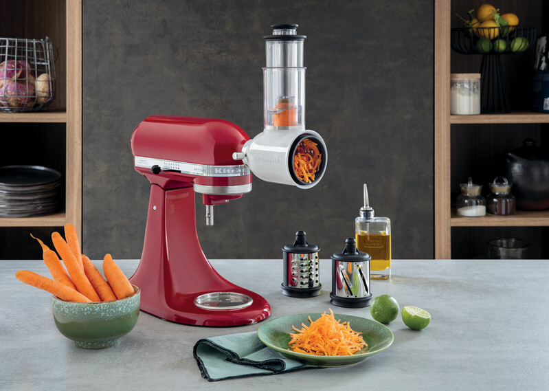 Red KitchenAid stand mixer with a grater attachment grating a carrot. Bowl of carrots and grated carrots on a plate.