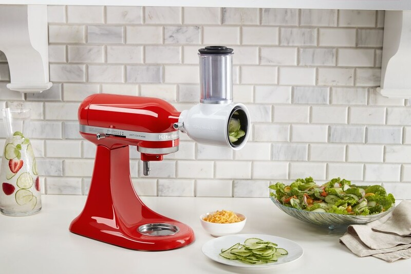 Red KitchenAid with slicer, slicing cucumbers. Salad and shredded cheese are nearby. Background is a white brick wall.