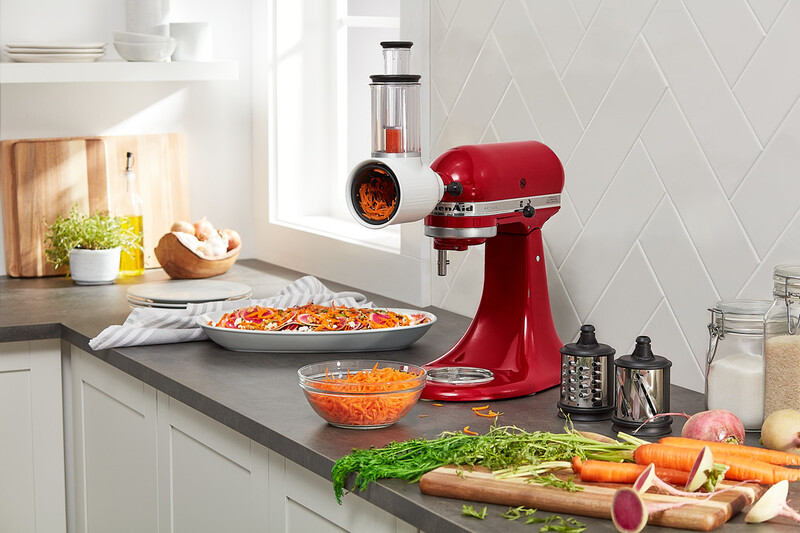 A red KitchenAid stand mixer with a shredder attachment. Carrots and other vegetables are prepared on a counter top.