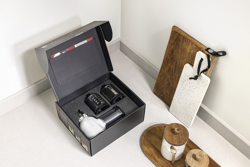 A box contains a food processor. Wooden and white cutting boards, a wooden tray with jars, and a white corner wall are present.