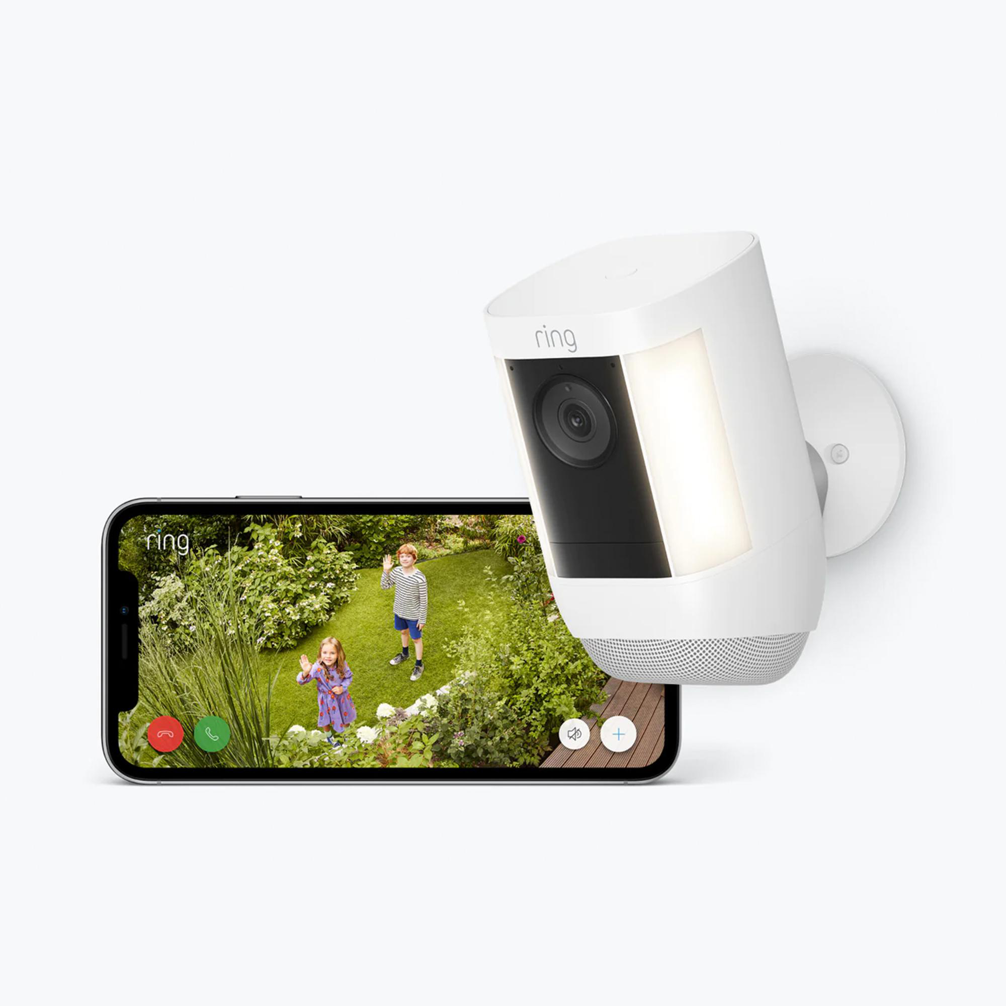 Ring-Kamera mit Telefon, das zwei Kinder in einem Garten zeigt. Kamera ist weiß, an der Wand befestigt. Telefon hat grüne Ruftaste.