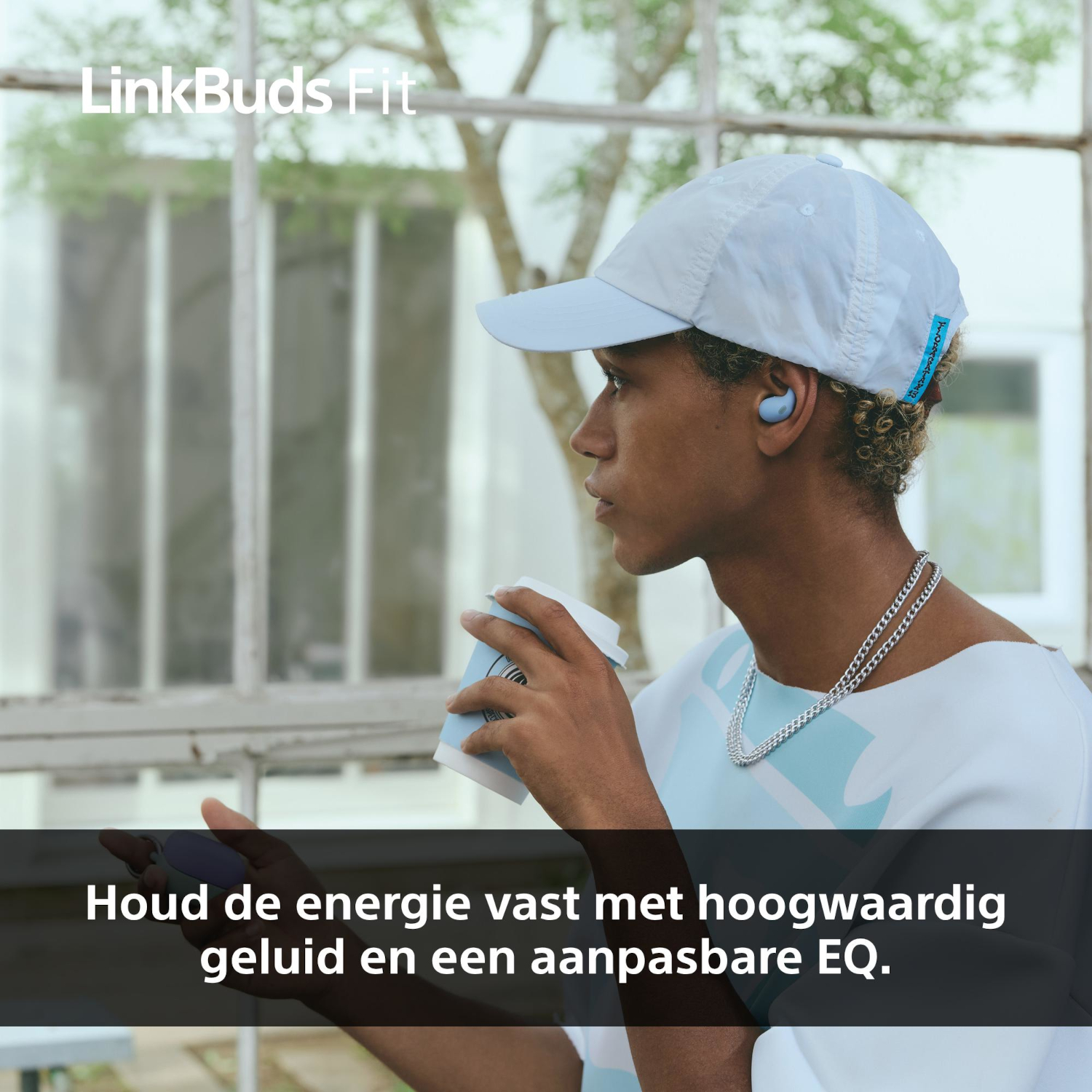 Een persoon met een blauwe pet en oortjes, die een koffiekopje vasthoudt, met een raam en bomen op de achtergrond.