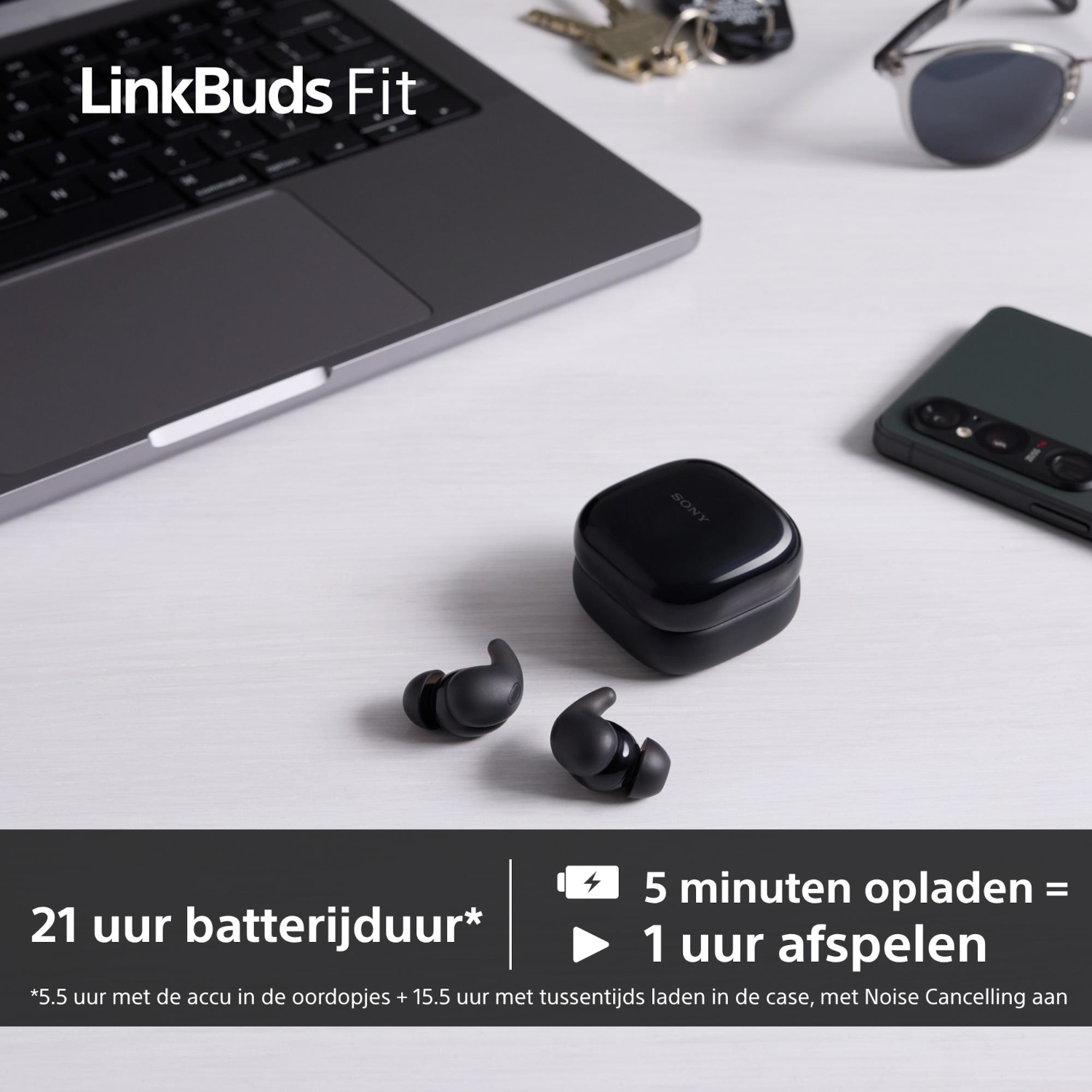 Zwarte Sony oordopjes en oplaadcase op een witte tafel. Een laptop, telefoon, zonnebril en sleutels zijn ook aanwezig. Tekst: 21h batterij.