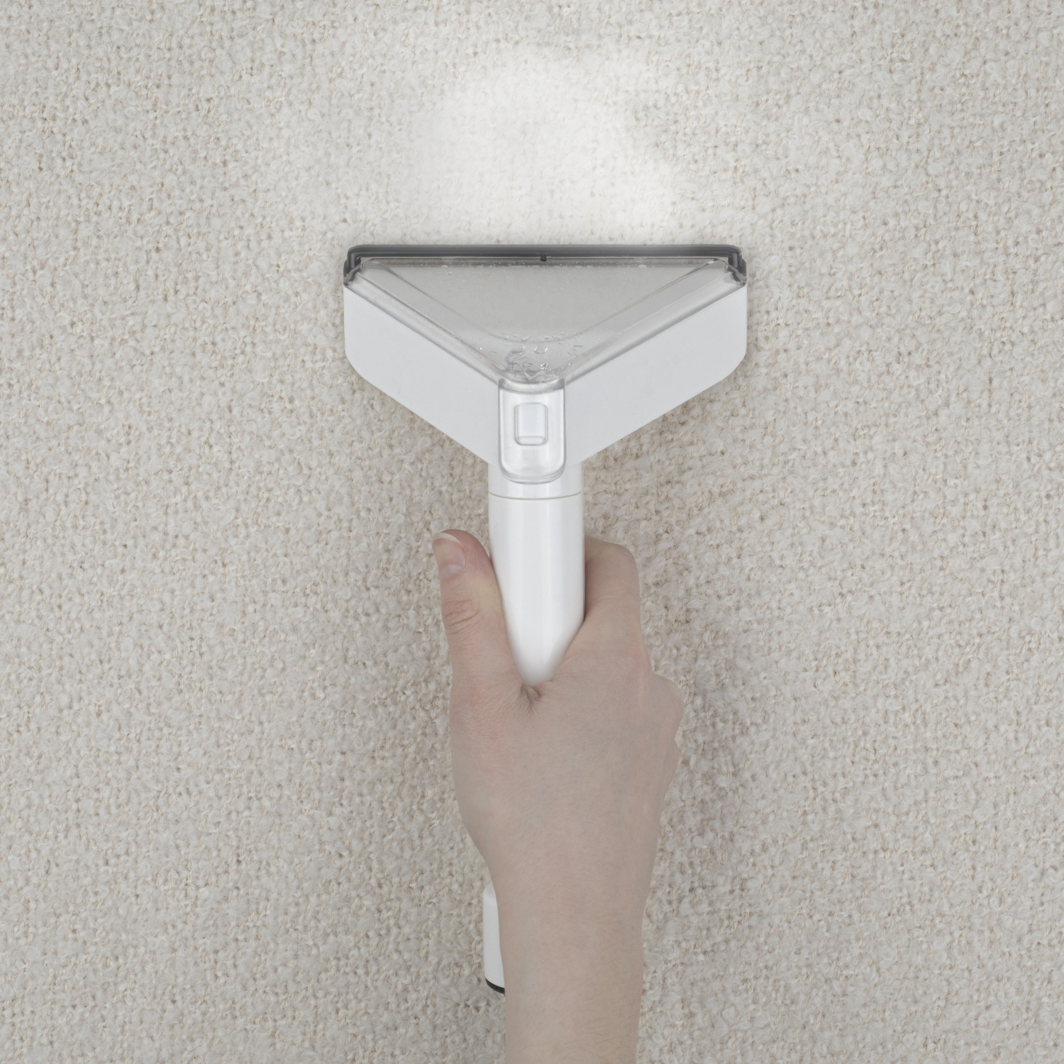 Una mano tiene un pulitore per tappeti bianco sopra un tappeto beige. La testina è triangolare, erogando vapore.