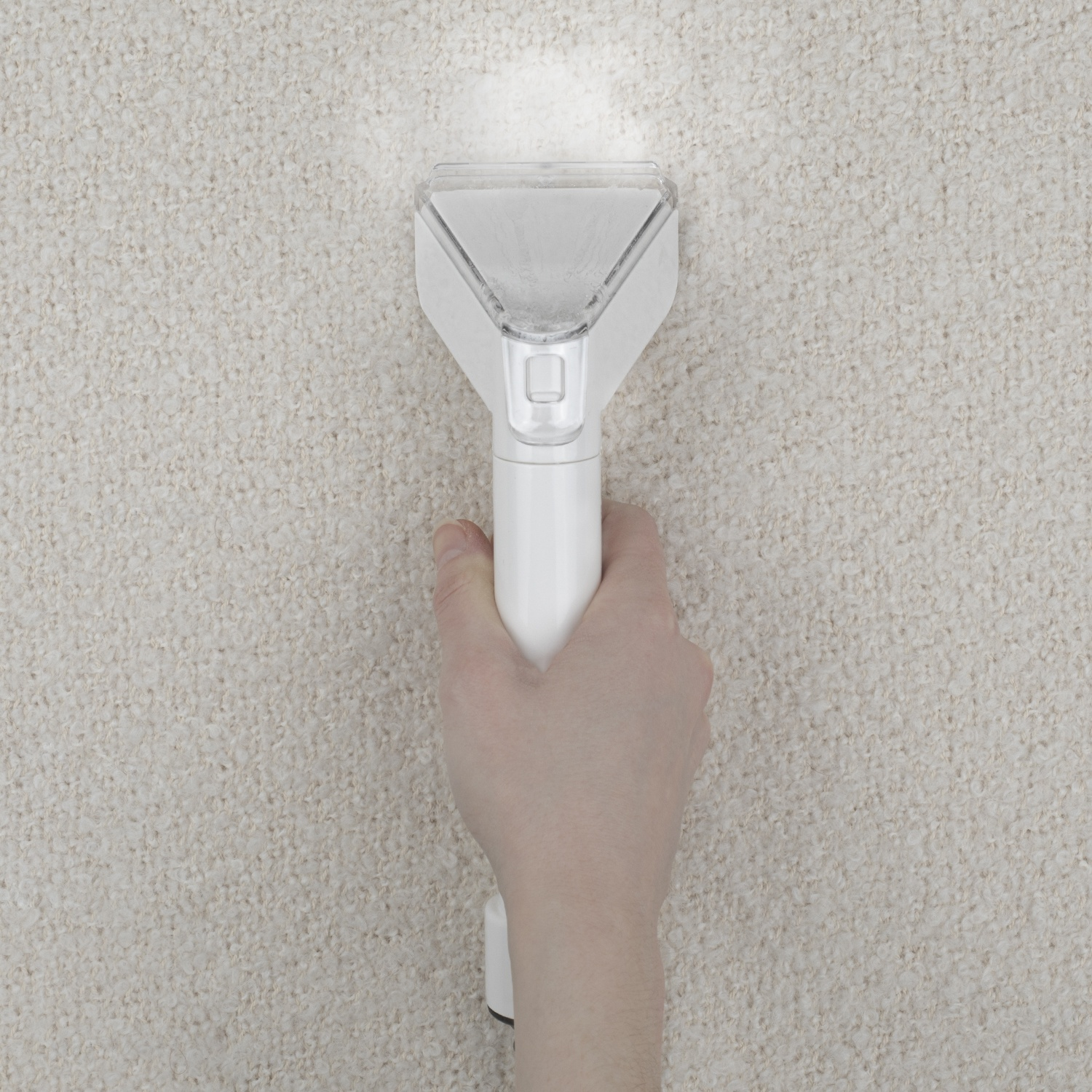 Una mano tiene un pulitore per tappeti bianco sopra un tappeto beige. La testa del pulitore è triangolare, la luce la illumina.