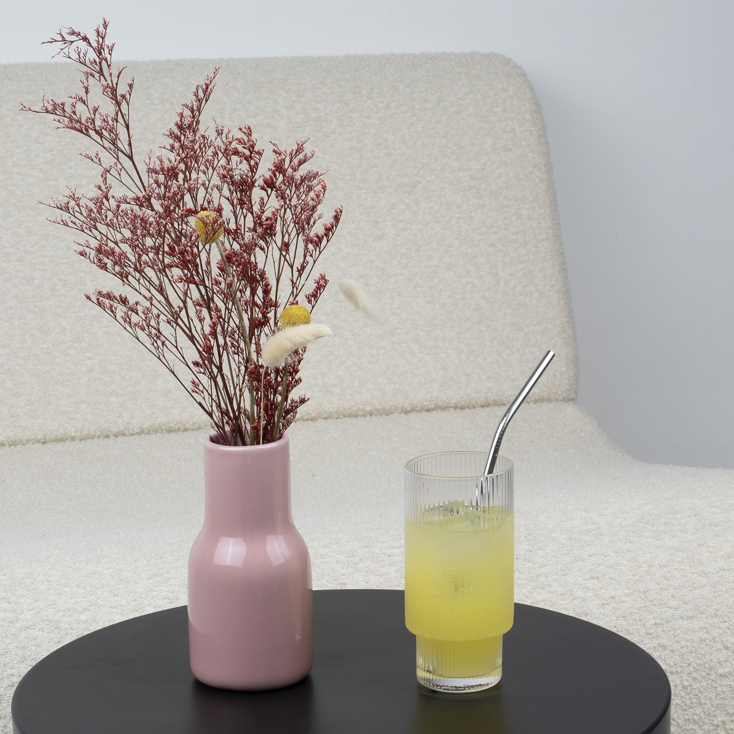 Un vaso rosa con fiori secchi su un tavolo nero. Un bicchiere di bevanda gialla con cannuccia. Una sedia bianca sullo sfondo.