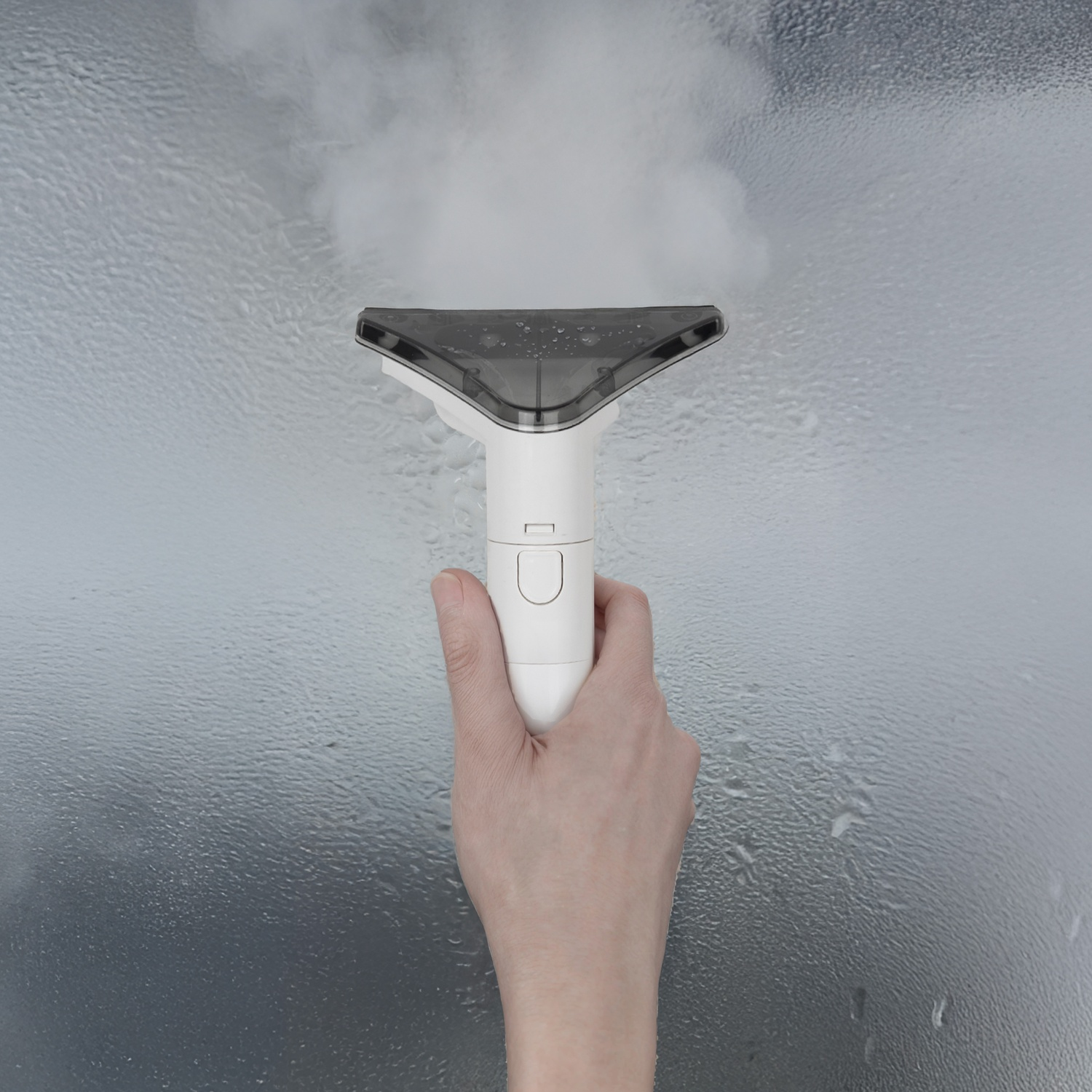Una mano tiene un pulitore a vapore bianco contro una finestra bagnata. Il vapore viene emesso, pulendo il vetro. Lo sfondo è un grigio sfocato.