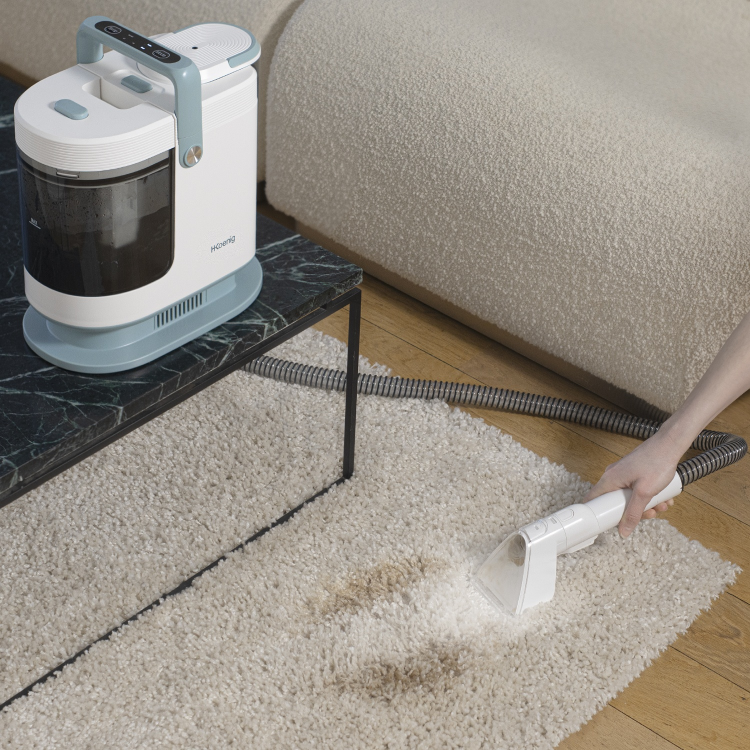 Un pulitore per tappeti bianco e turchese viene utilizzato su un tappeto beige macchiato. Una mano tiene il pulitore. Un divano bianco e un tavolo sono sullo sfondo.