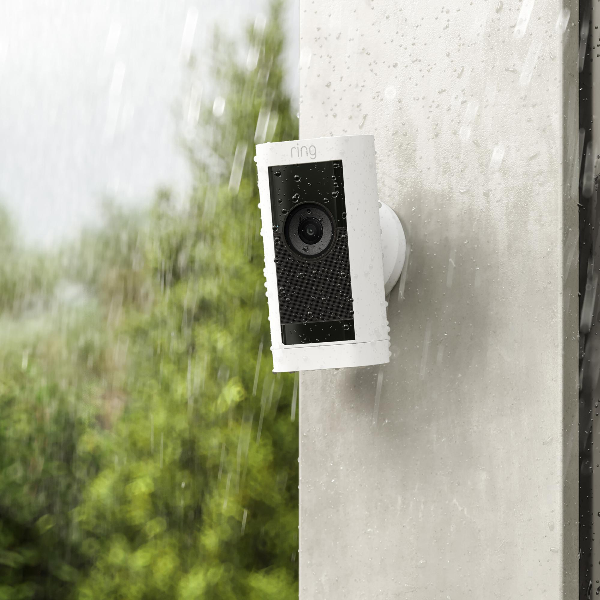 Eine weiße Ring-Überwachungskamera ist im Regen an einer Wand montiert. Grünes Laub im unscharfen Hintergrund.