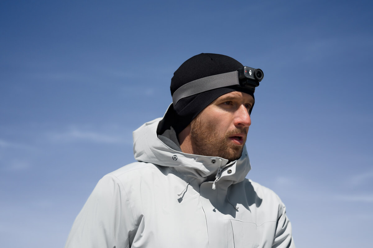 Mann mit schwarzer Mütze, grauem Riemen mit Kamera, hellgrauer Jacke und Blick zur Seite vor blauem Himmel.