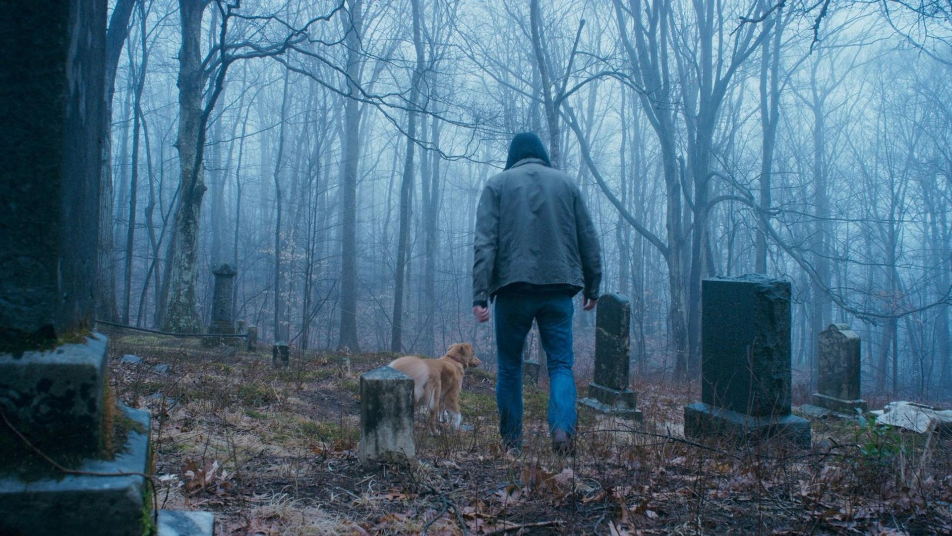 Ein Mann und ein Hund gehen durch einen nebligen Friedhof. Kahle Bäume umgeben die Gegend mit sichtbaren Grabsteinen.