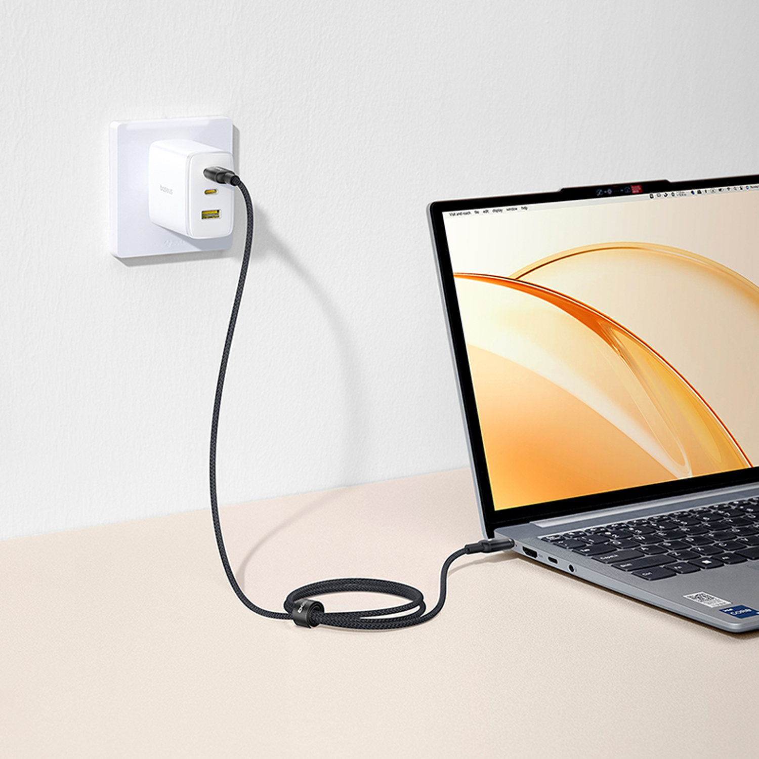Cargando portátil con cable negro. Cargador blanco enchufado a la pared. Portátil en superficie beige, fondo blanco.