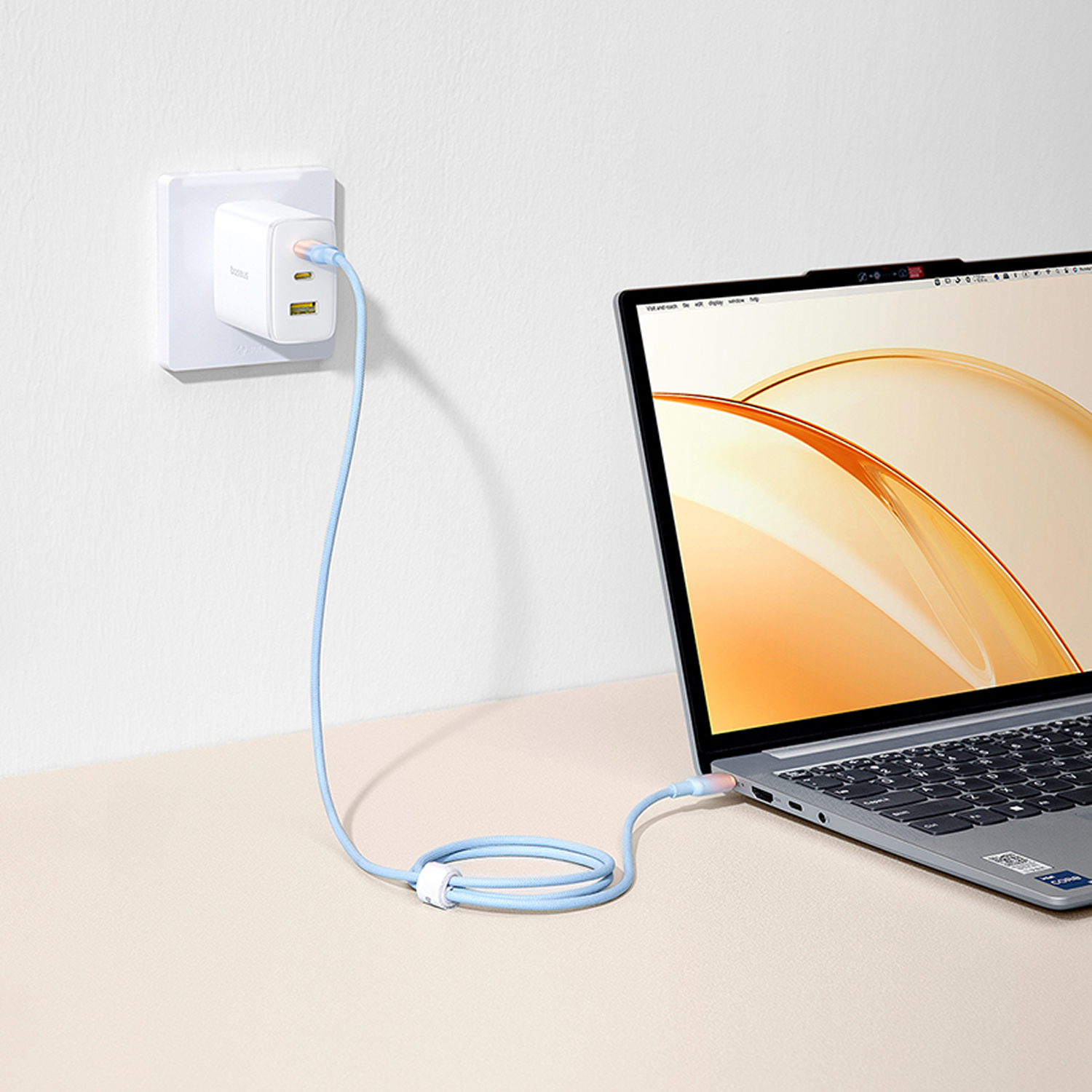 Portátil cargándose con un cable azul. El cargador está enchufado a una toma de corriente. El portátil está sobre una superficie beige.