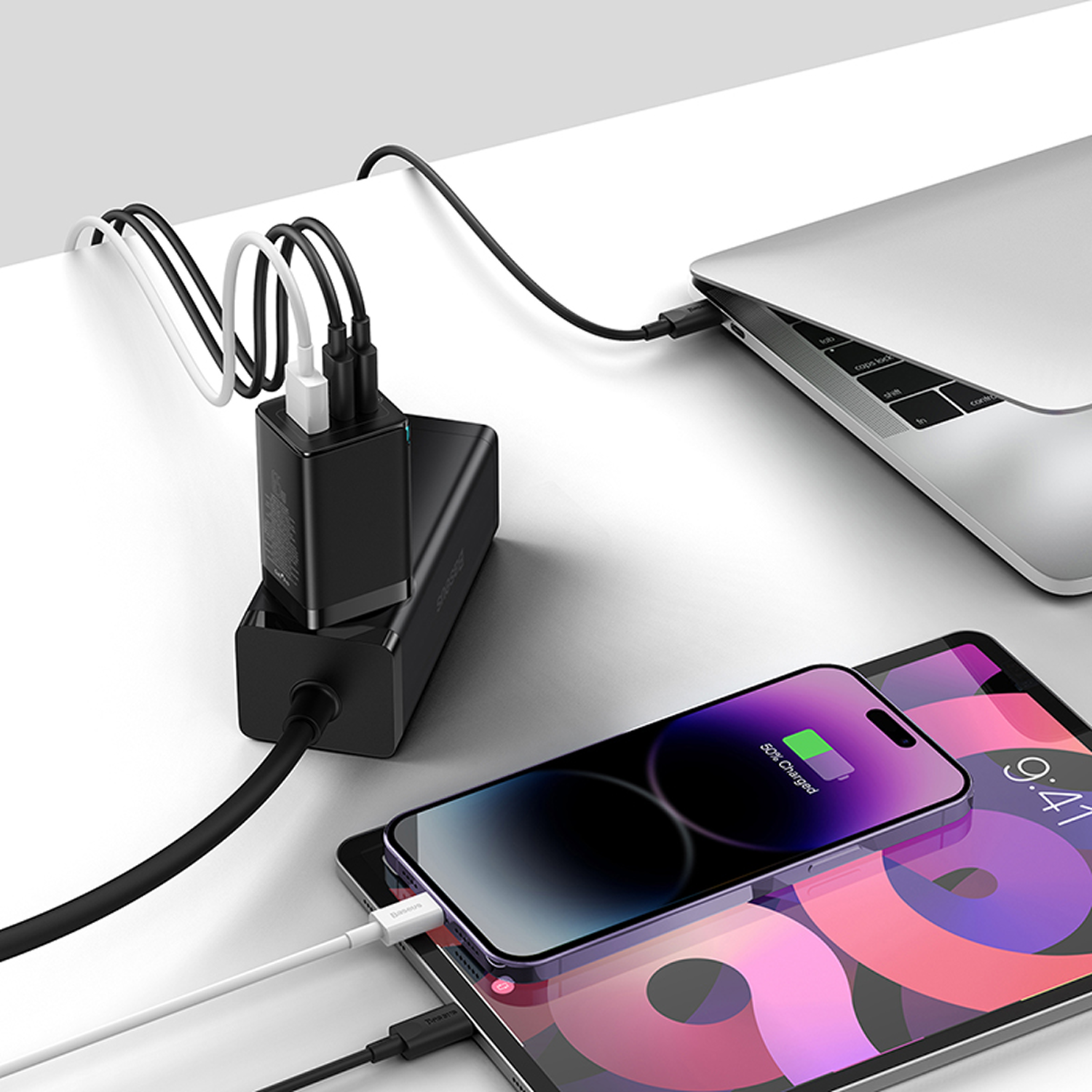 Un portátil, una tableta y un teléfono se cargan con un cargador multipuerto. El teléfono muestra un 50% de carga. Los cables son negros y blancos.