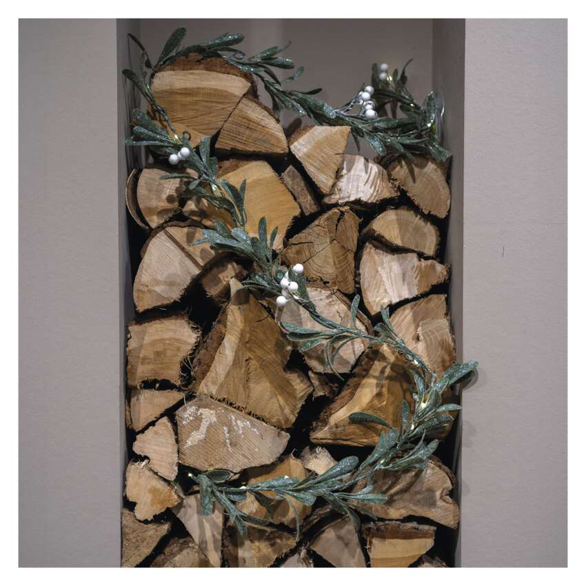 Egy farakás zöld levelekből és fehér bogyókból álló girlanddal van díszítve. A fa egy süllyesztett falban van.