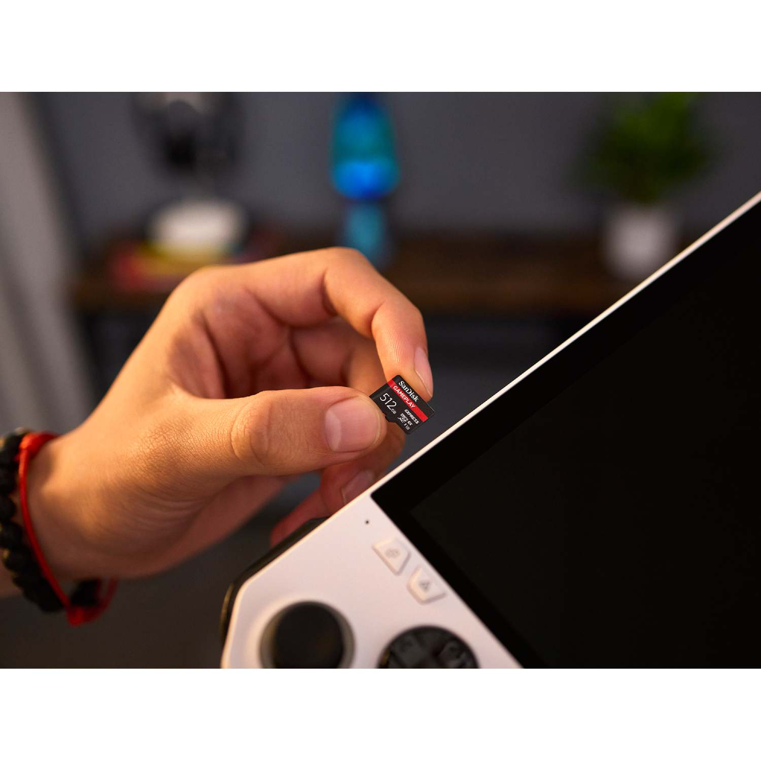 Una mano sostiene una tarjeta microSD SanDisk de 512GB cerca de un dispositivo de juegos portátil blanco. El fondo tiene una estantería de madera borrosa.