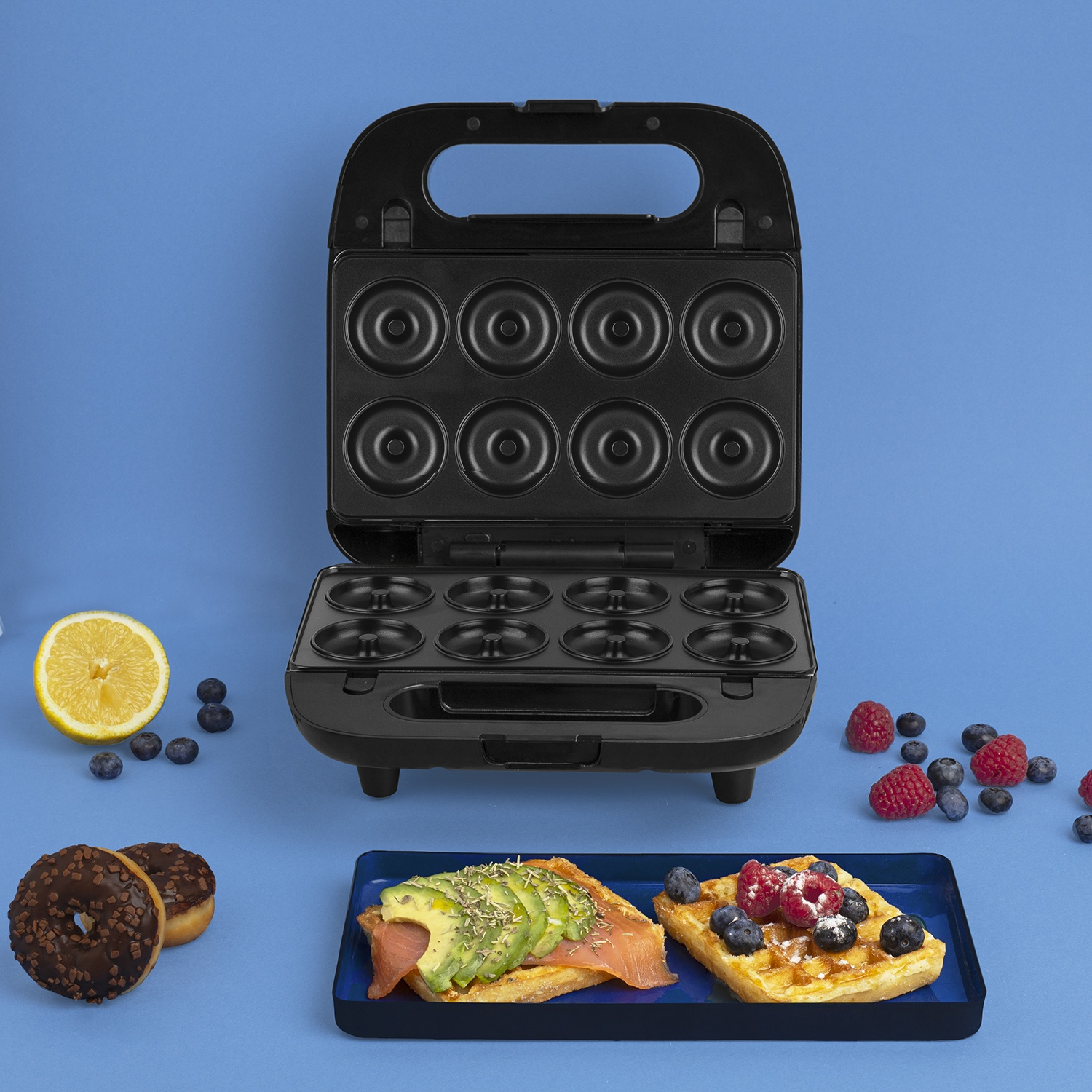Una macchina per fare ciambelle nera, waffle, ciambelle, bacche, toast con avocado e limone su una superficie blu.