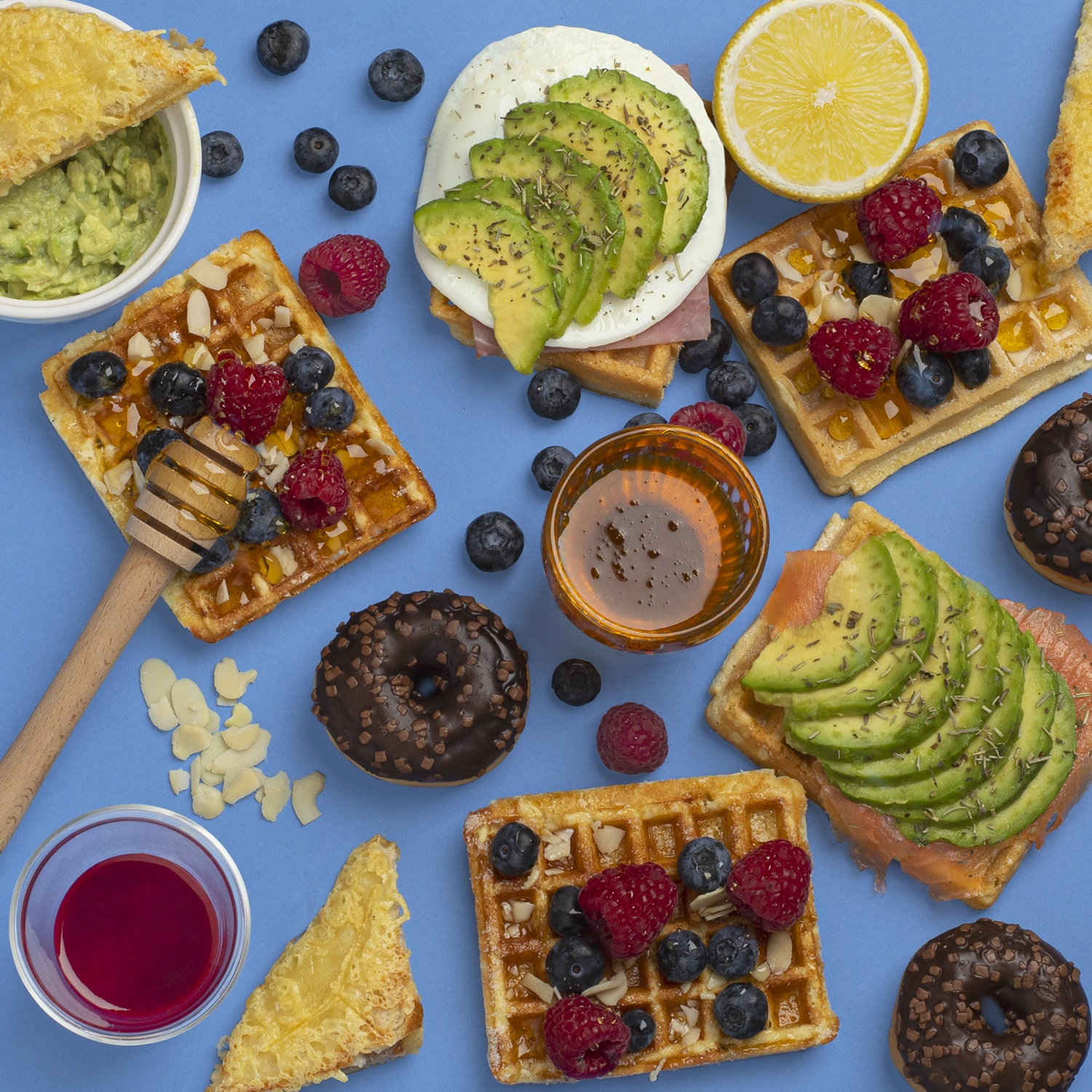 Vista dall'alto di waffle, toast con avocado, bacche, ciambelle, miele e limone su sfondo blu.