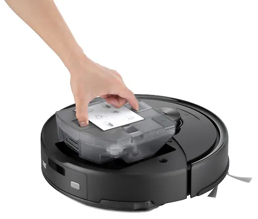 Egy kéz átlátszó tartályt helyez egy fekete robotporszívóra. A tartályban fehér papír van utasításokkal.