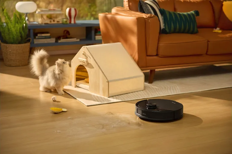 Egy fehér macska egy ház alakú macskaágyat néz. Egy robotporszívó tisztítja a fa padlót egy barna kanapé közelében.