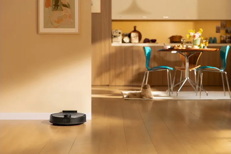 Robotporszívó a fapadlón. Egy macska pihen a háttérben. Konyha és étkező látható.
