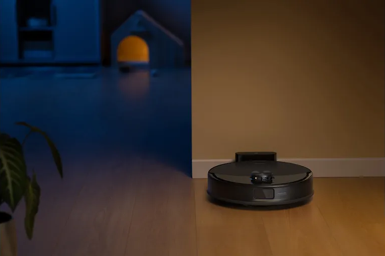 Egy fekete robot porszívó fa padlón, a fal közelében. A háttérben egy ház alakú tárgy sárga fénnyel.