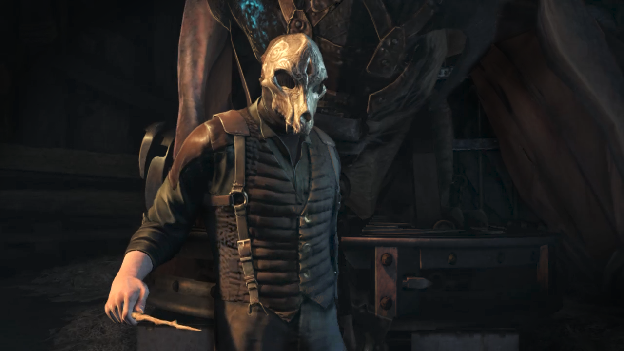 Mężczyzna w masce czaszki i ciemnym stroju trzyma mały patyk. Duża postać wisi za nim obok metalowego stołu.