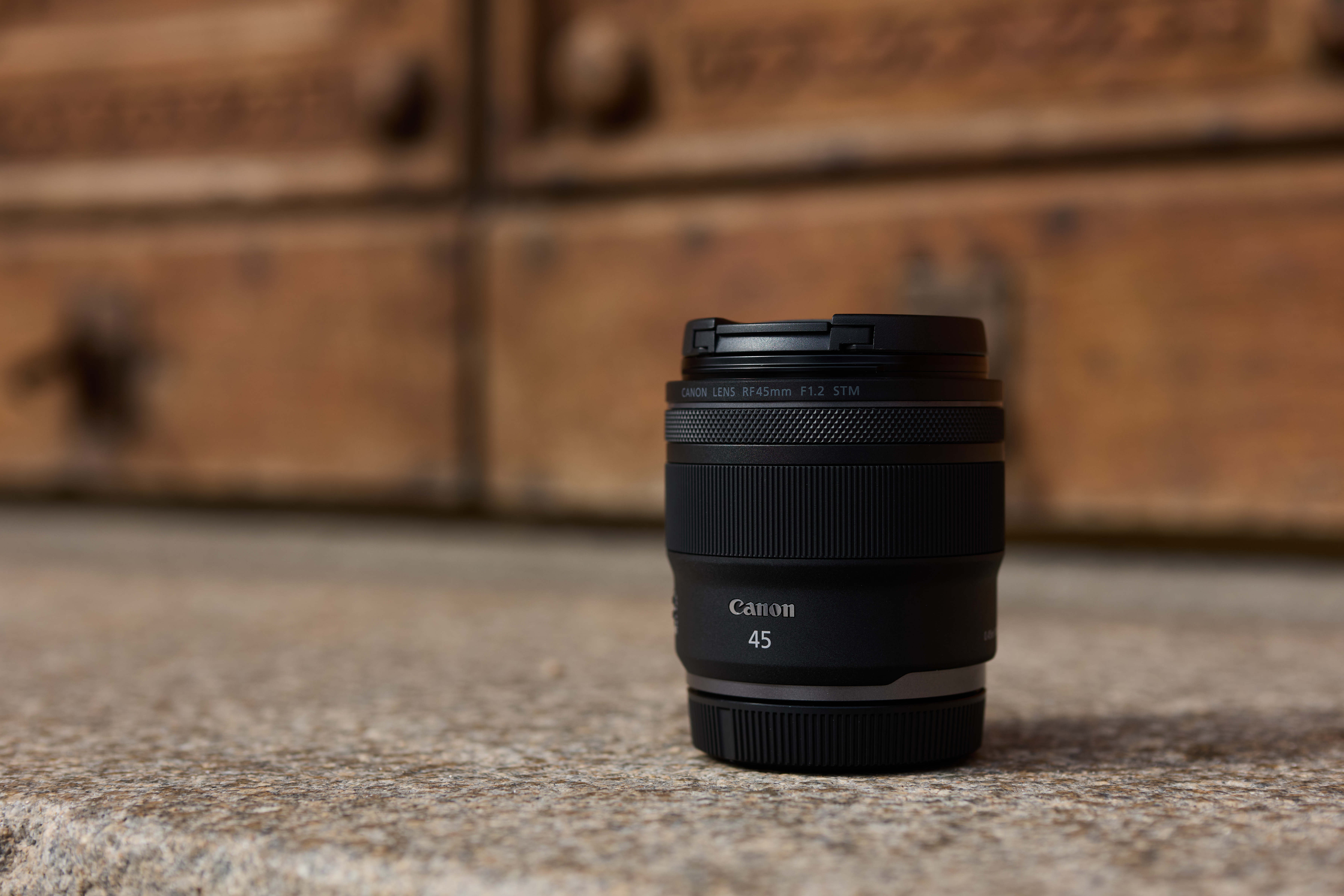 Ein schwarzes Canon 45mm Objektiv liegt auf einer Steinoberfläche. Der Hintergrund ist eine verschwommene Holztür.