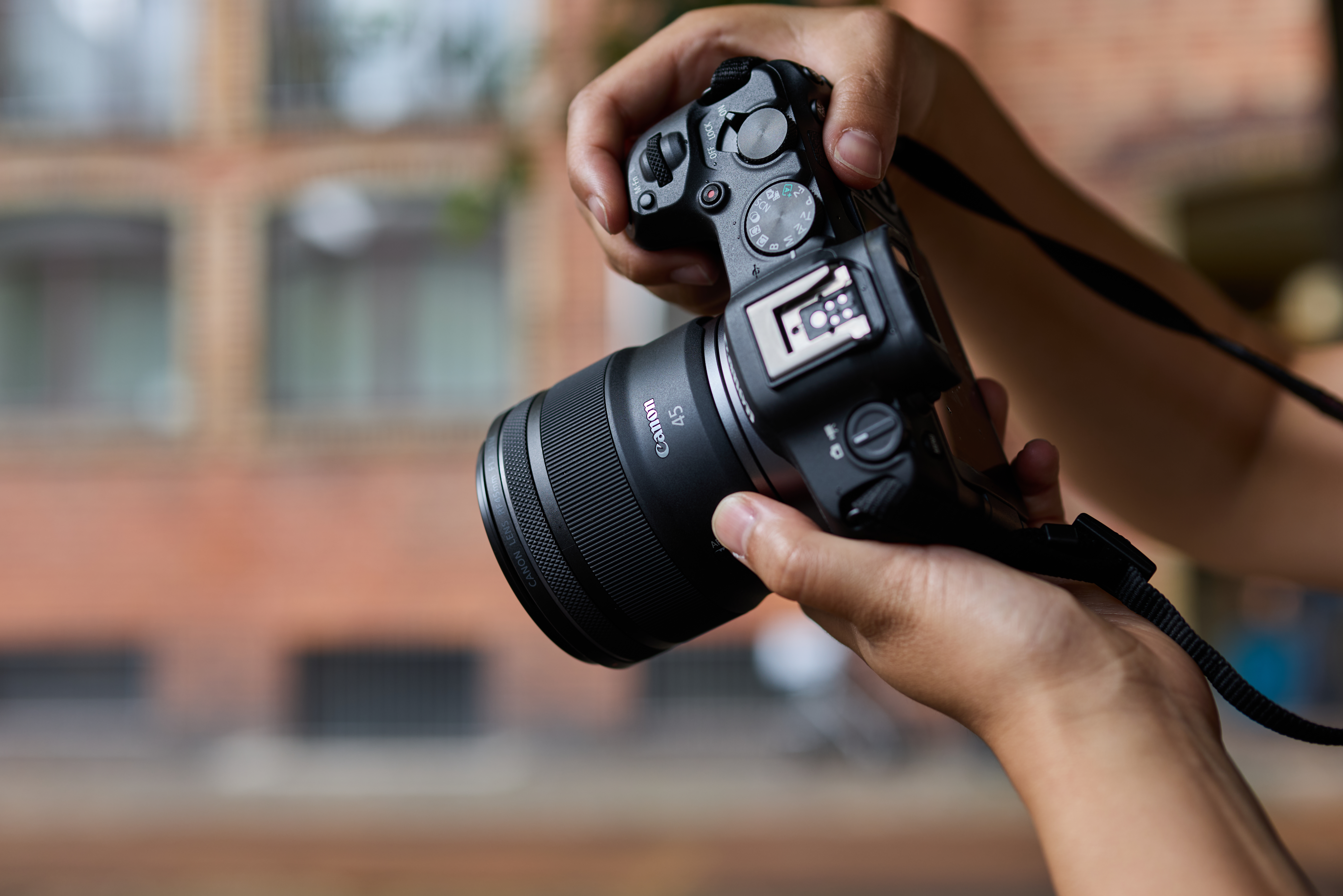 Eine Person hält eine Canon-Kamera mit einem 45-mm-Objektiv. Ein Backsteingebäude ist im Hintergrund unscharf.