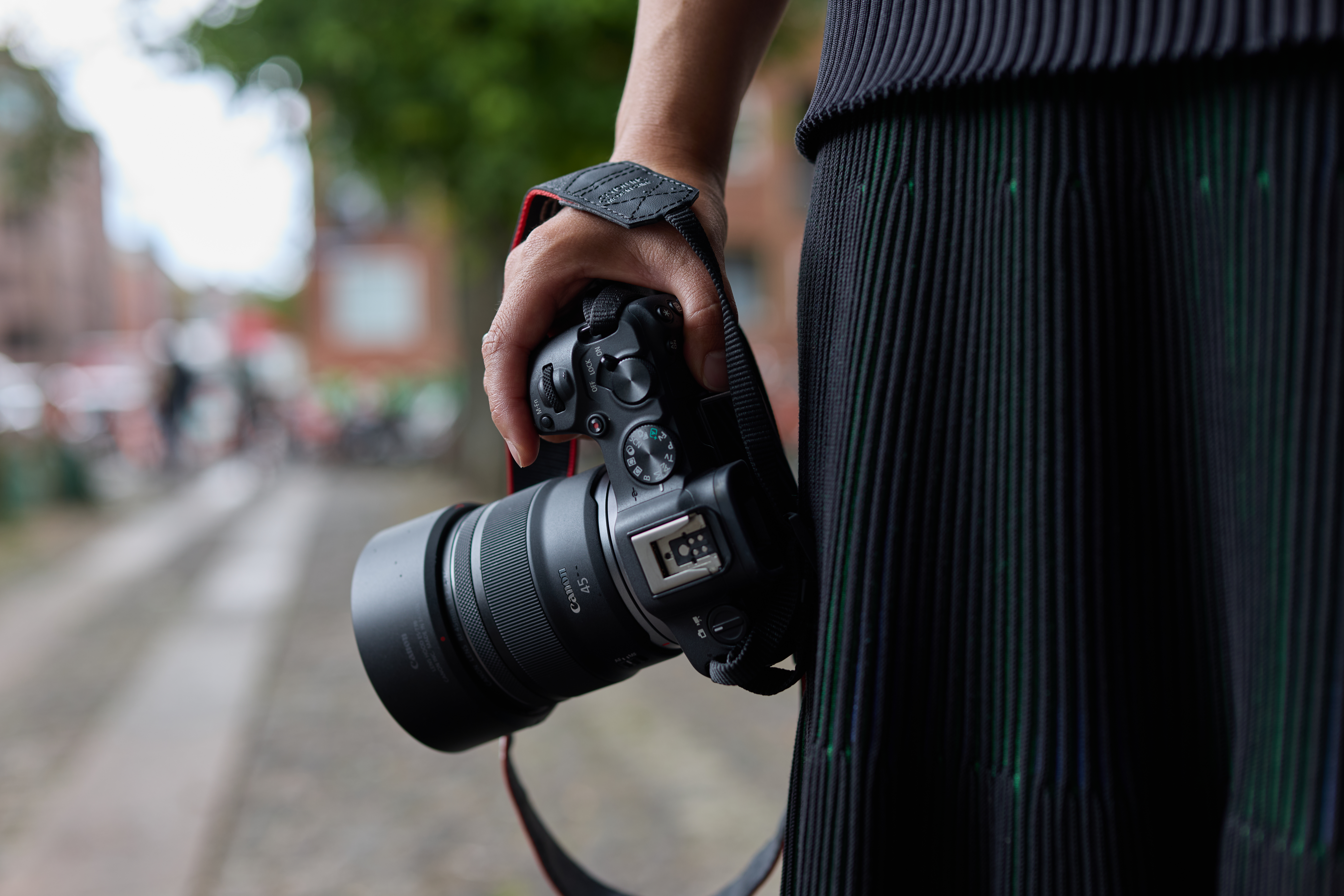 Eine Person hält eine Canon-Kamera. Das Objektiv zeigt nach links. Ein schwarzes Kleid befindet sich rechts. Der Hintergrund ist unscharf.