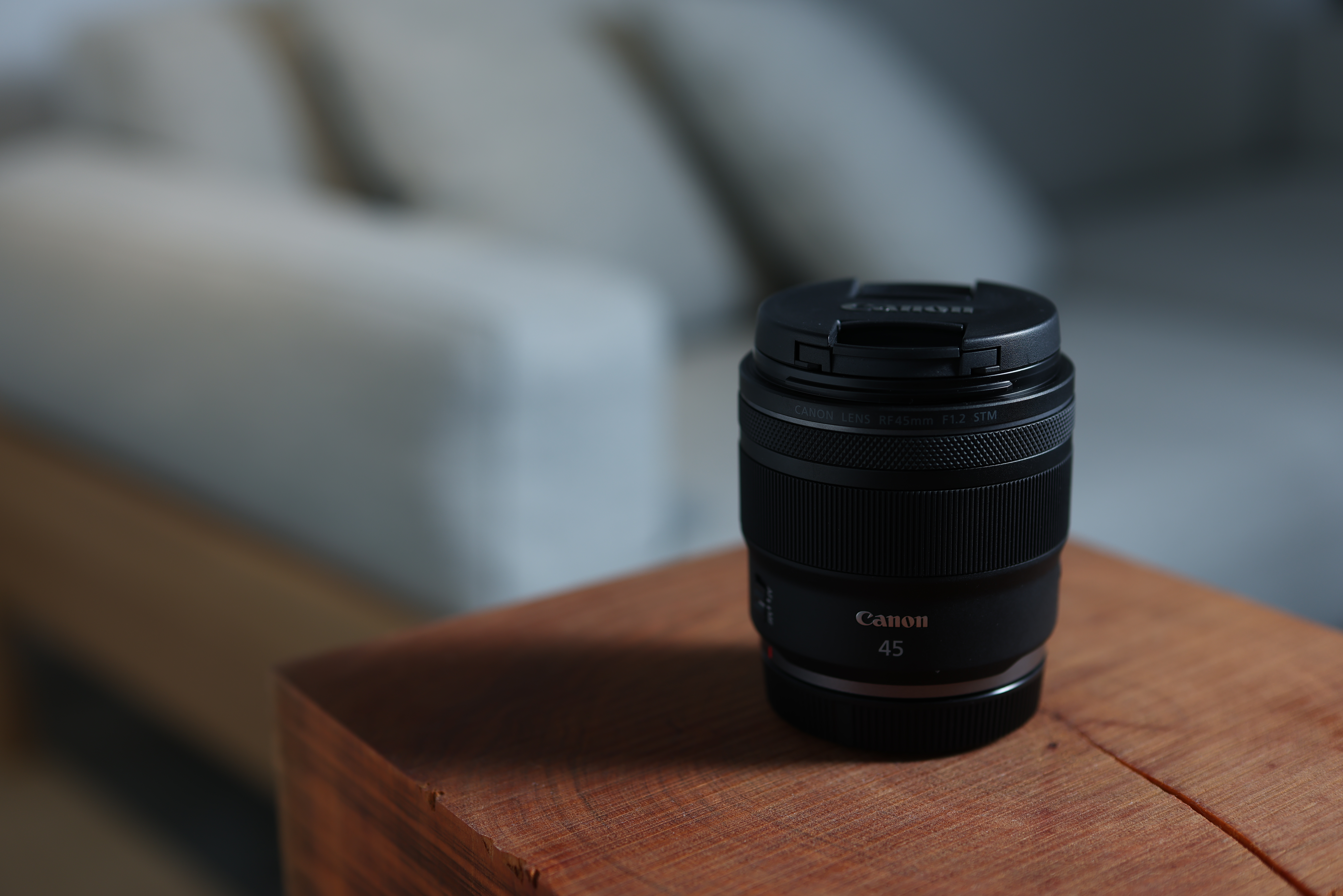 Ein Canon RF 45mm F1.2 STM-Objektiv mit Objektivdeckel steht auf einer Holzoberfläche, unscharfer Hintergrund.