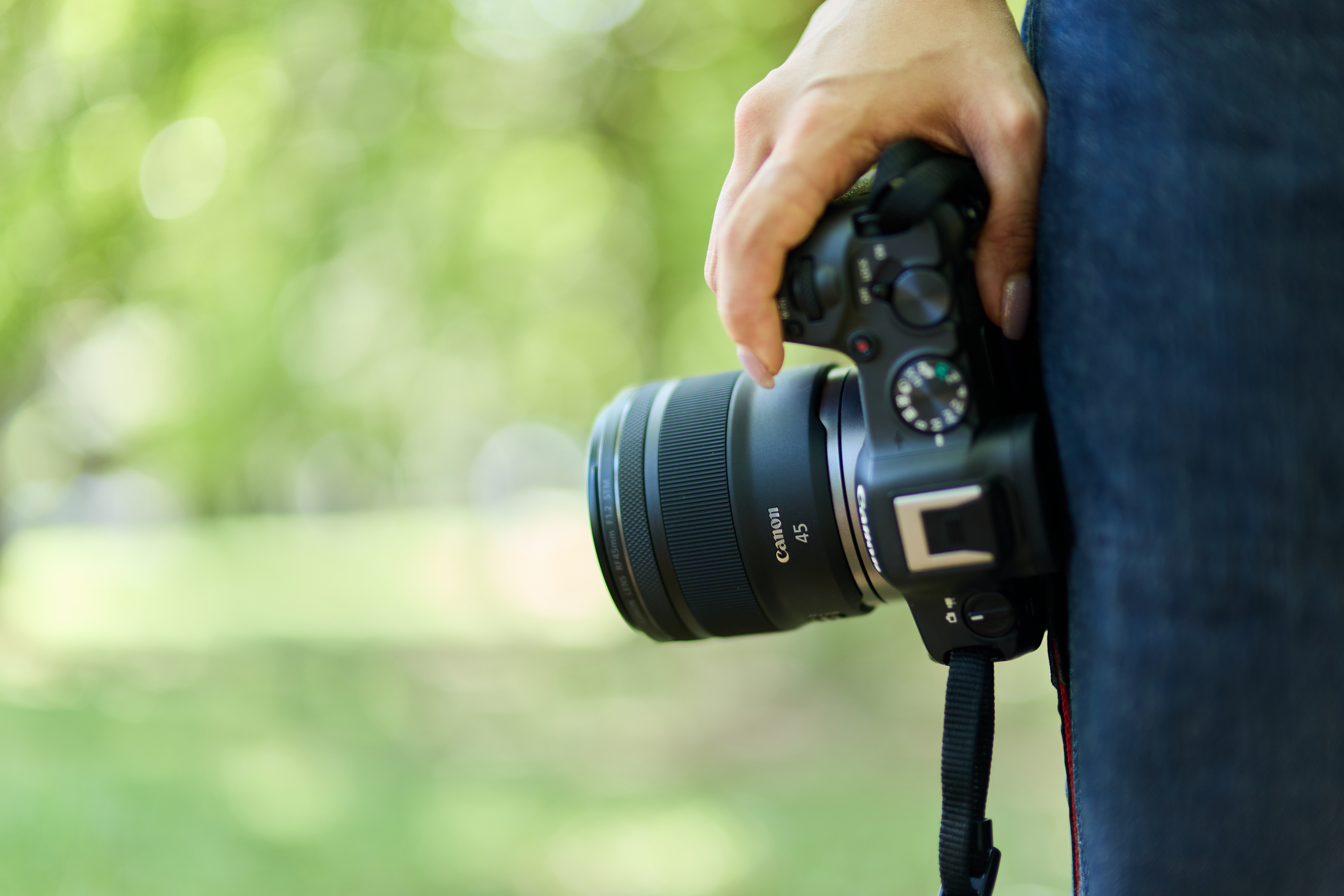 Eine Person hält eine Canon-Kamera mit einem 45-mm-Objektiv. Grüner Bokeh-Hintergrund. Die Person trägt Jeans.