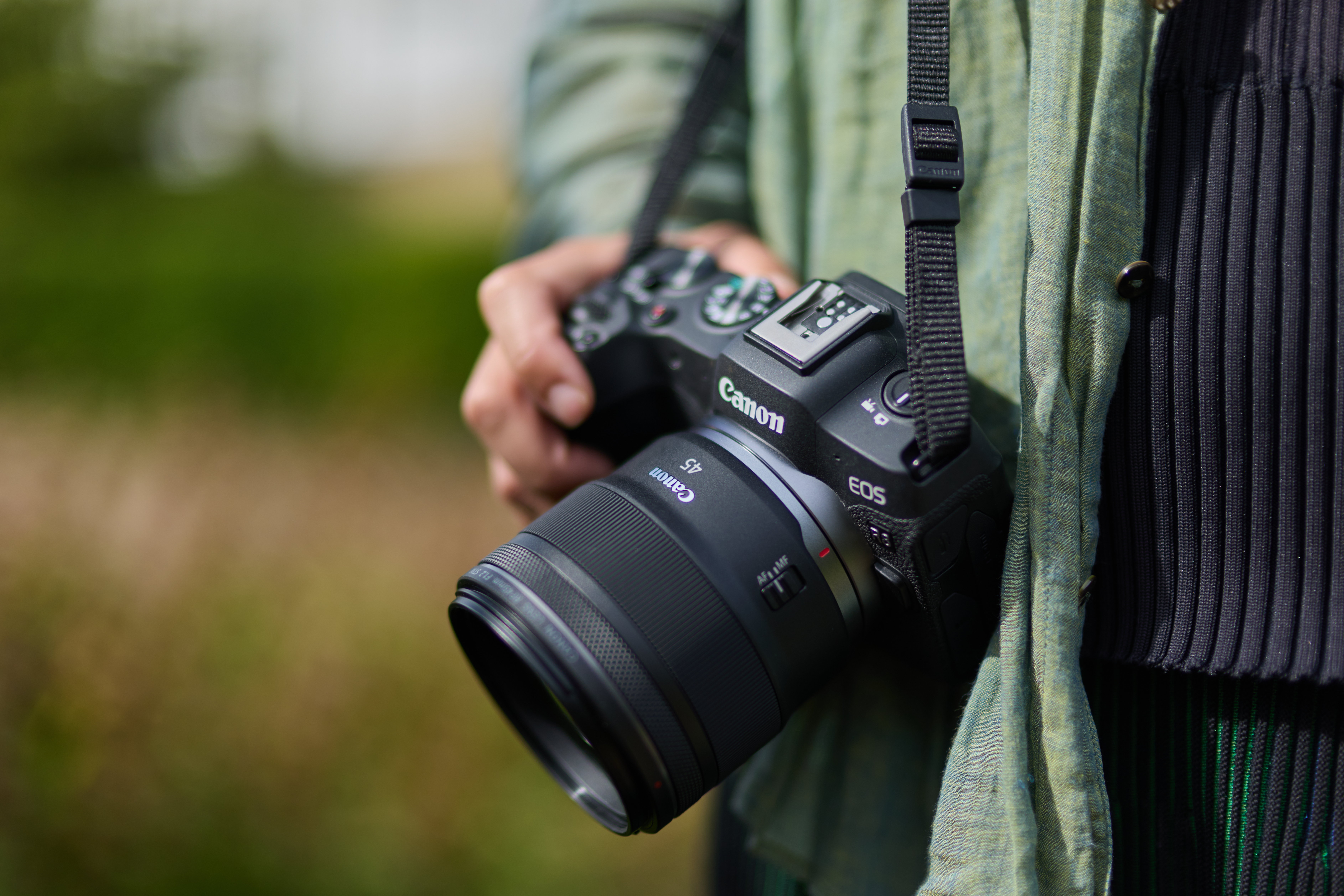 Eine Person, die eine Canon EOS-Kamera hält. Der Kameragurt ist schwarz. Die Person trägt ein grünes Hemd.