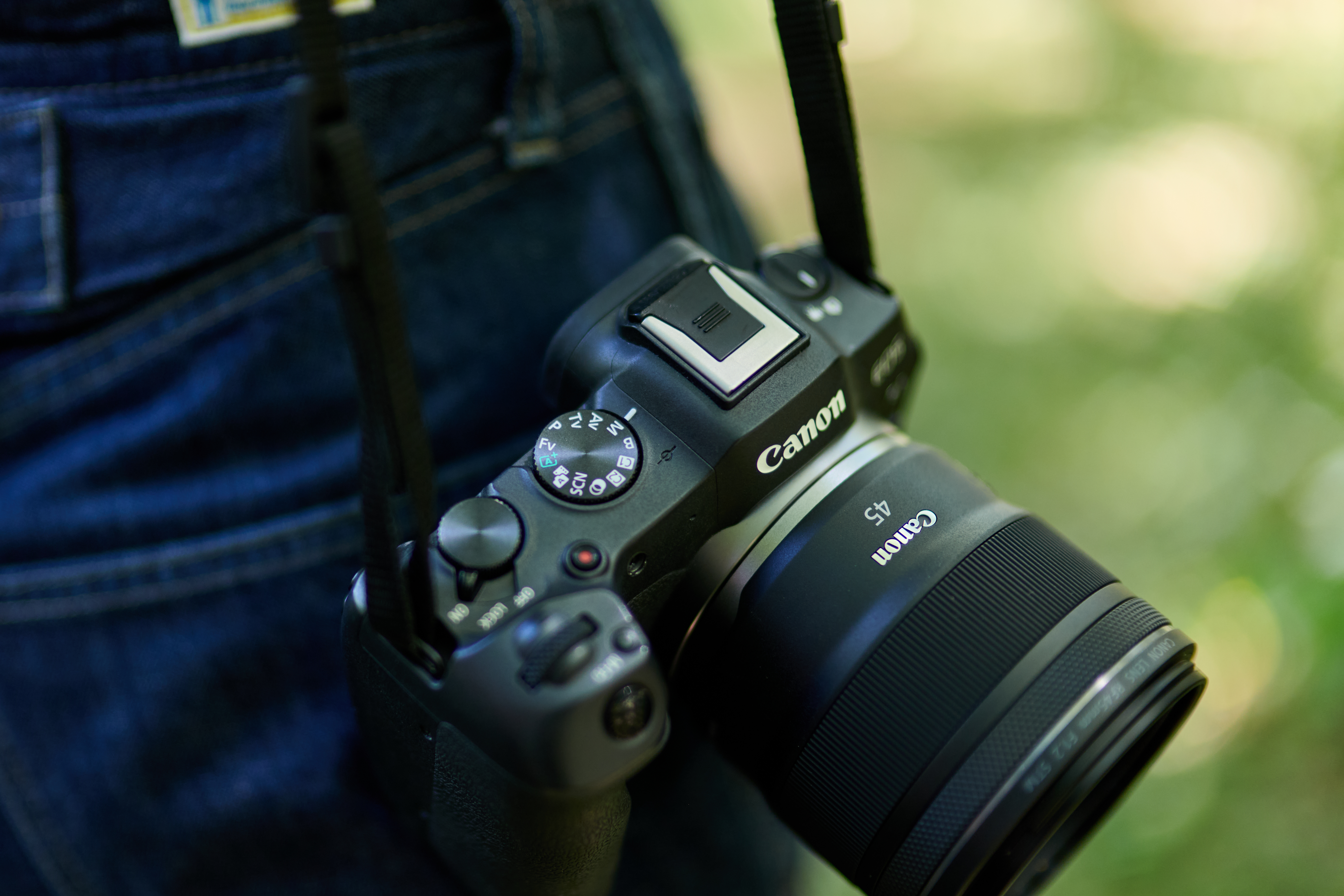 Eine schwarze Canon-Kamera mit 45-mm-Objektiv hängt. Sie liegt an einer Person mit Jeans vor unscharfem grünem Hintergrund.