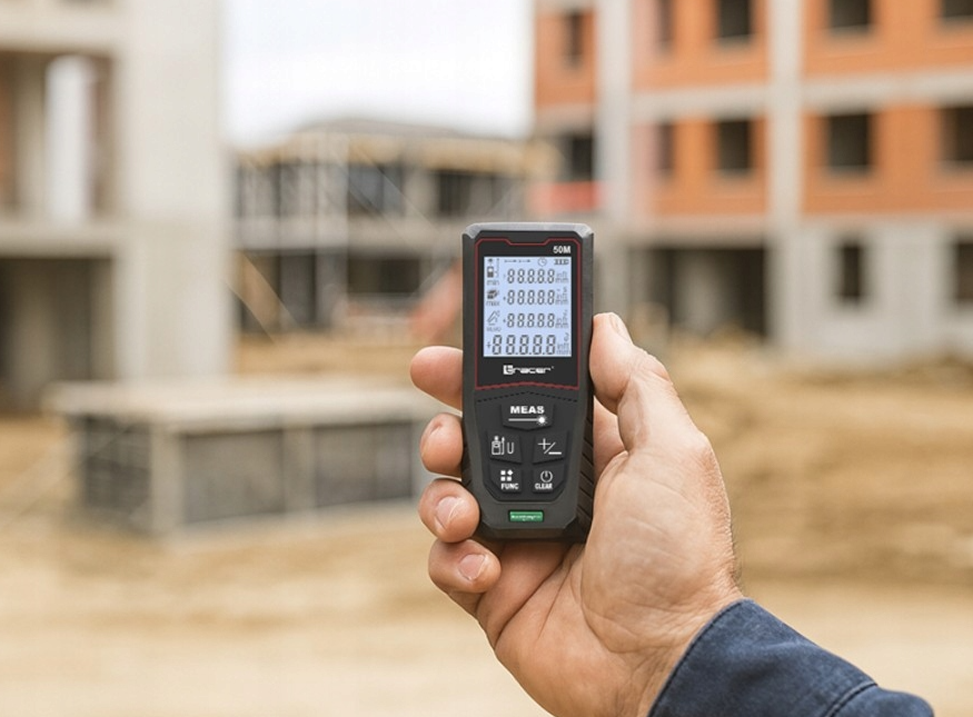 Ręka trzyma czarno-czerwony laserowy miernik odległości, w tle budynek w budowie.