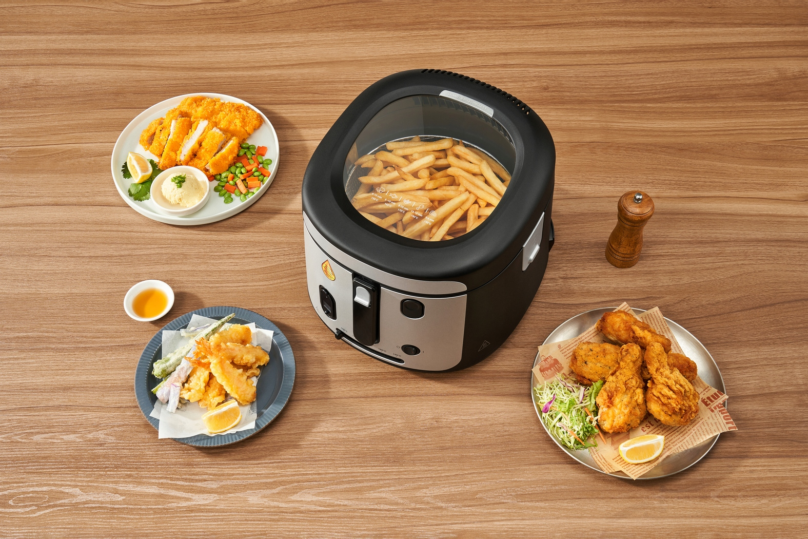 Widok z góry na smażone potrawy: kurczak, frytki, tempura i kotlety. Frytownica stoi na środku. Stół jest drewniany.