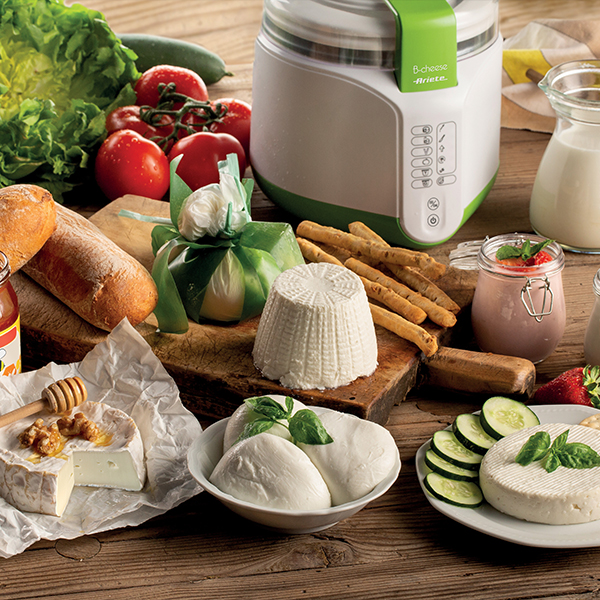 Na drewnianym stole wystawione są różne sery, chleb, pomidory i serowar. Jest słoik miodu i talerz z pokrojonymi ogórkami.