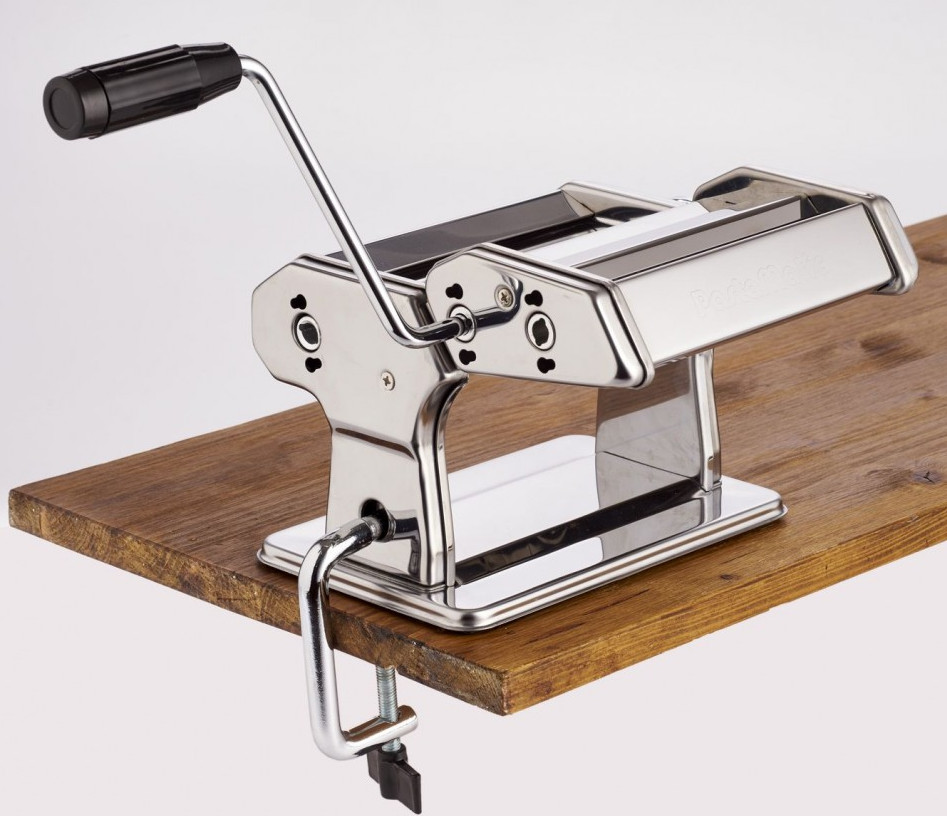 Maszyna do makaronu, srebrno-czarna, stoi na drewnianej powierzchni. Ma uchwyt i zacisk. Tło jest białe.