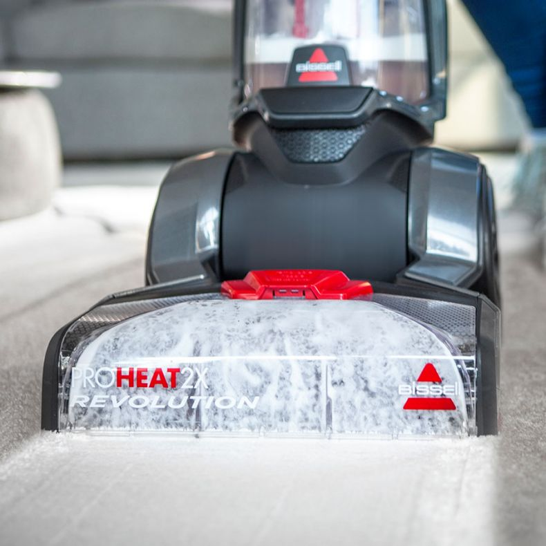 Odkurzacz do dywanów Bissell ProHeat 2X Revolution czyści jasny dywan. Widoczna jest piana i woda. Maszyna jest czarna i czerwona.